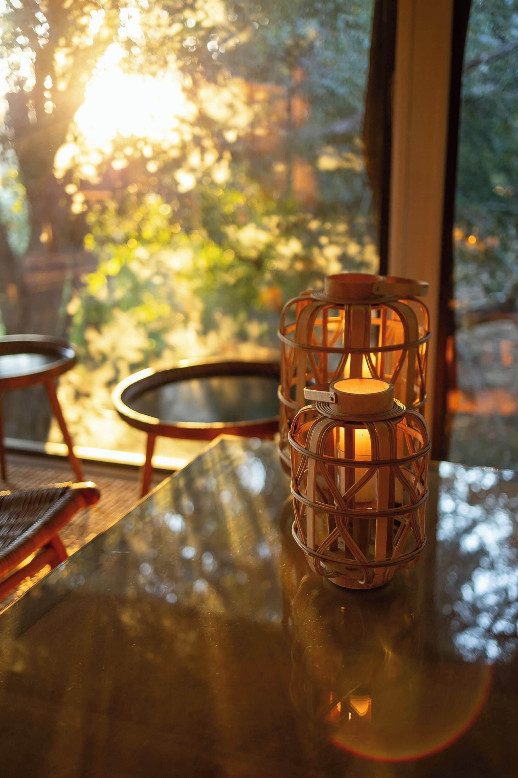 Una vela en un soporte tejido sobre una mesa de vidrio, con vista a árboles a través de una ventana.