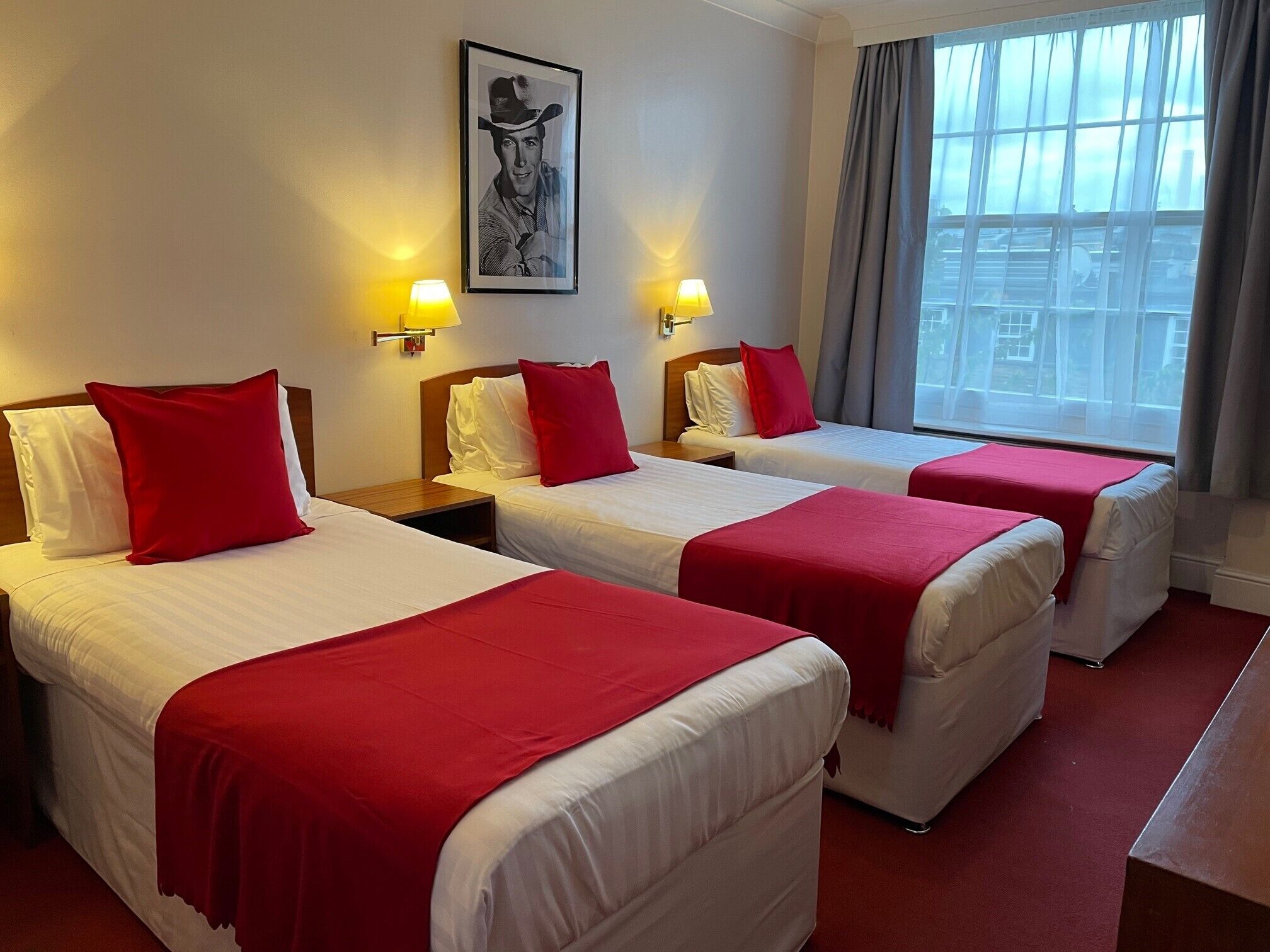 A hotel room with three beds, each with white bedding and red accents, a large window with a view of the city, and two wall-mounted lamps.