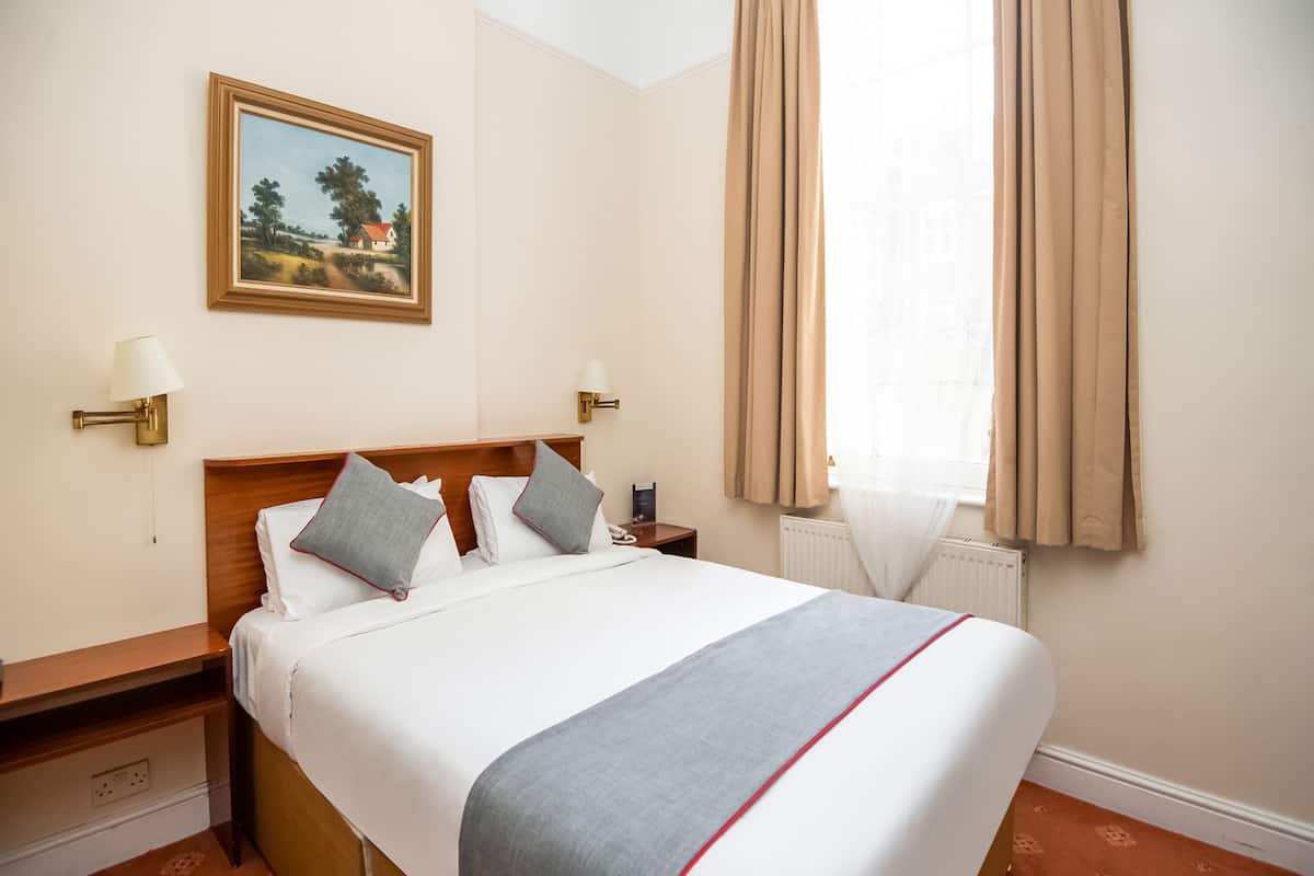 Habitación de hotel con una cama bien hecha, un cuadro en la pared, una mesita de noche con lámpara y una ventana con cortinas.