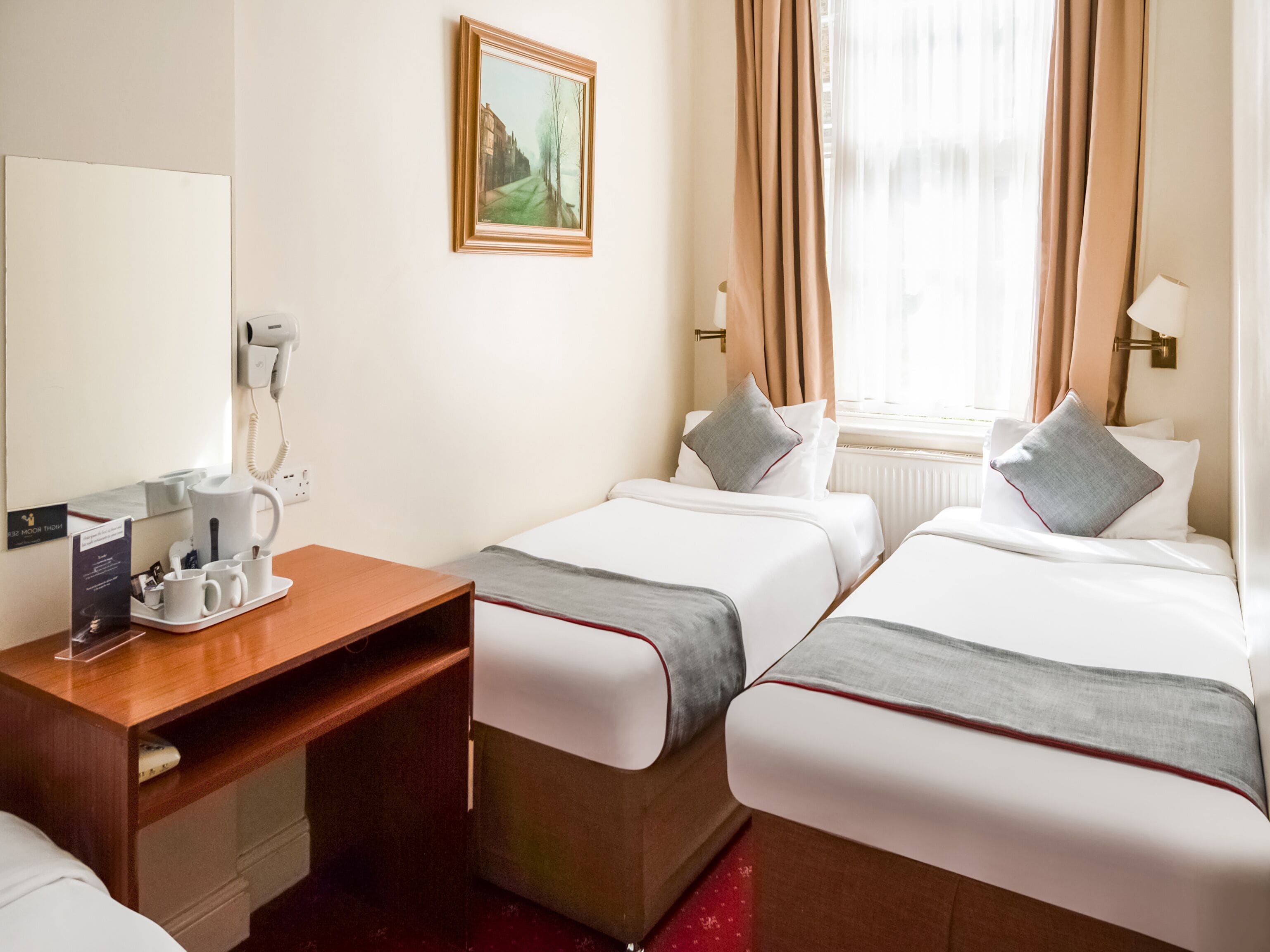 A hotel room with two beds, a desk with a telephone and coffee maker, a painting on the wall, and a window with curtains.