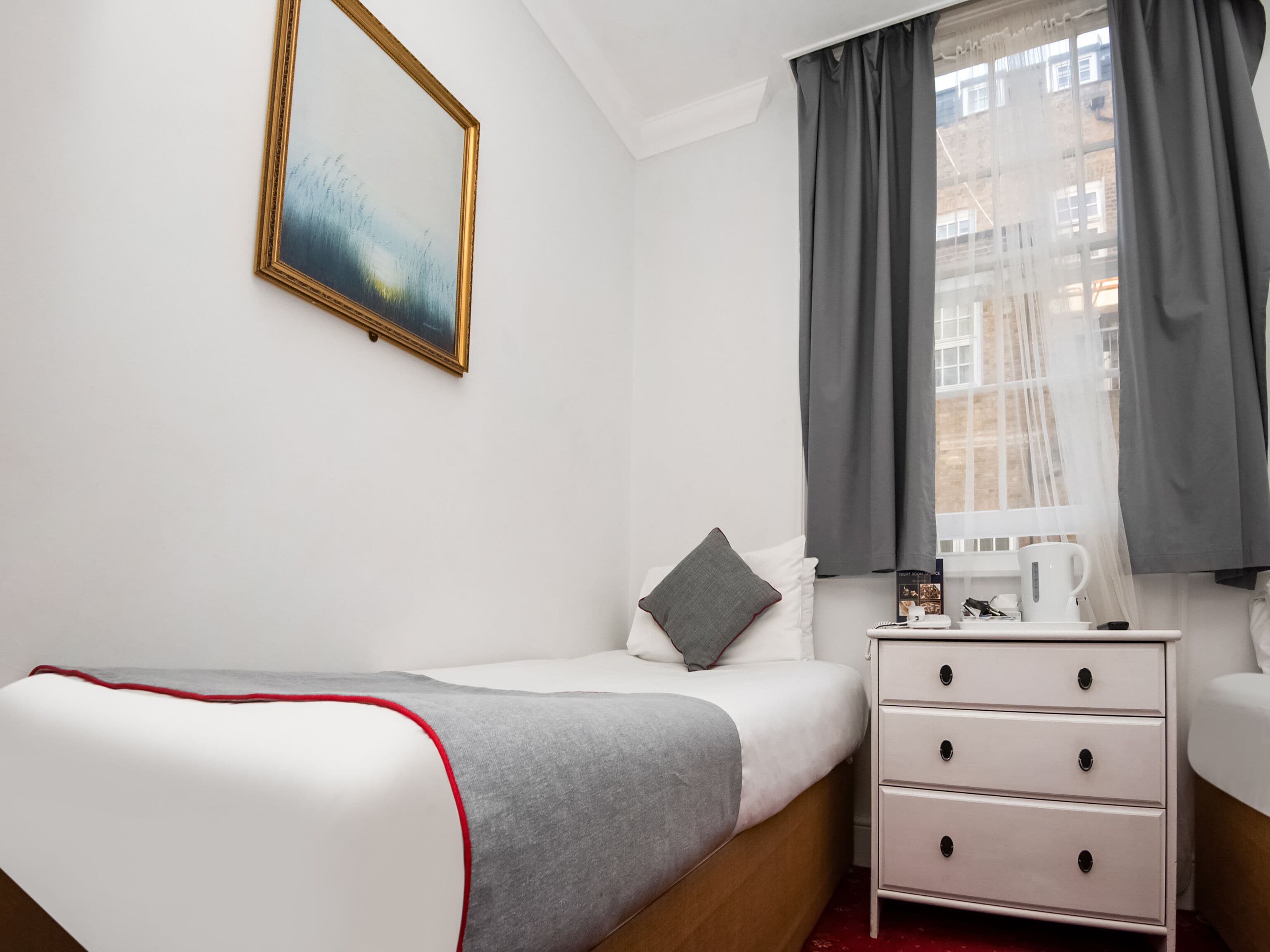 A hotel room with a single bed, a bedside table with a kettle, and a window with curtains.
