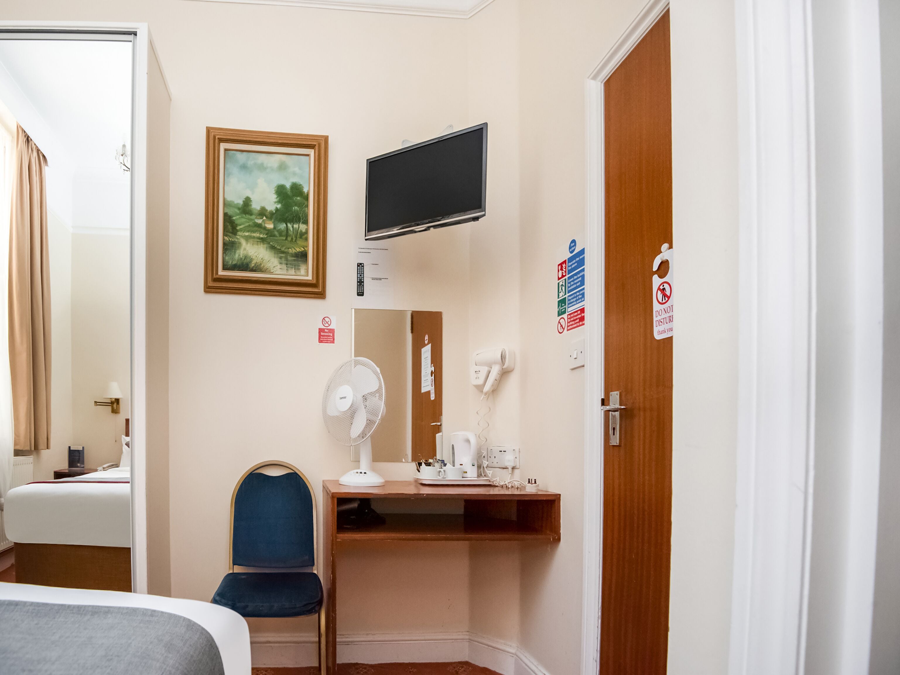 Una habitación de hotel con cama, escritorio con computadora, silla, ventilador, televisor y un cuadro en la pared.