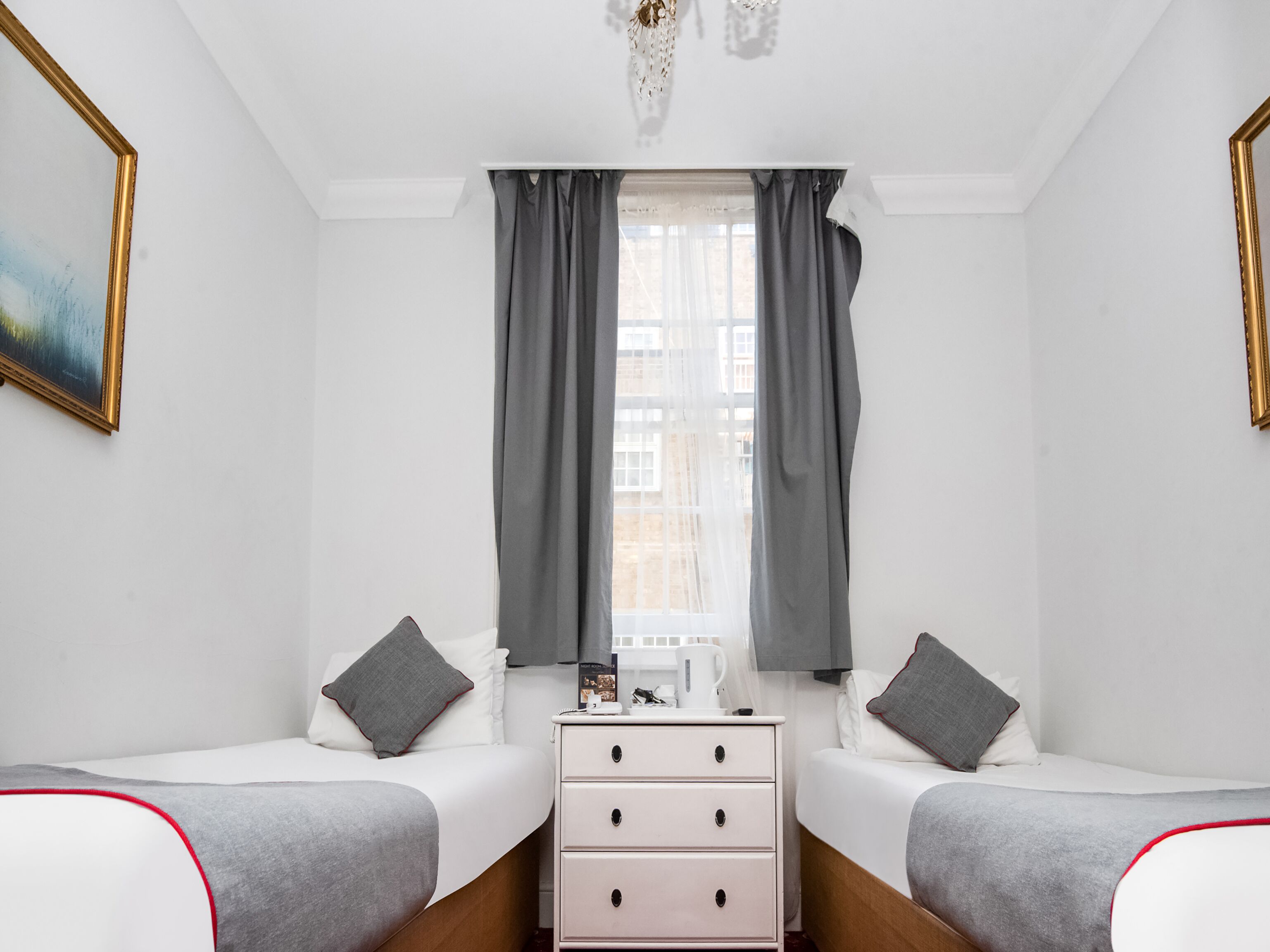 A hotel room with two beds, a dresser, a window with curtains, and a painting on the wall.