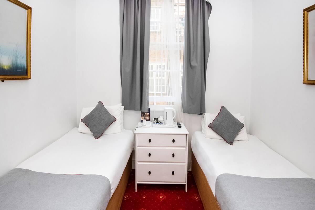 Una habitación de hotel con dos camas individuales, una mesita de noche con cafetera, una lámpara y una ventana con cortinas.