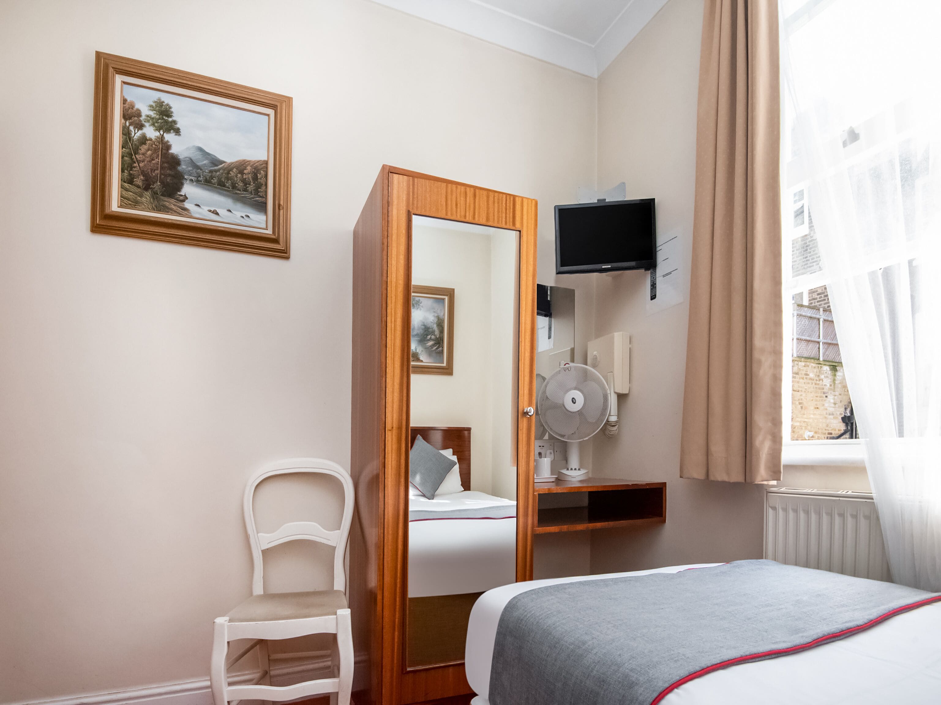 Une chambre d’hôtel avec un lit, une chaise, un bureau avec un ordinateur, une télévision et une fenêtre avec des rideaux.