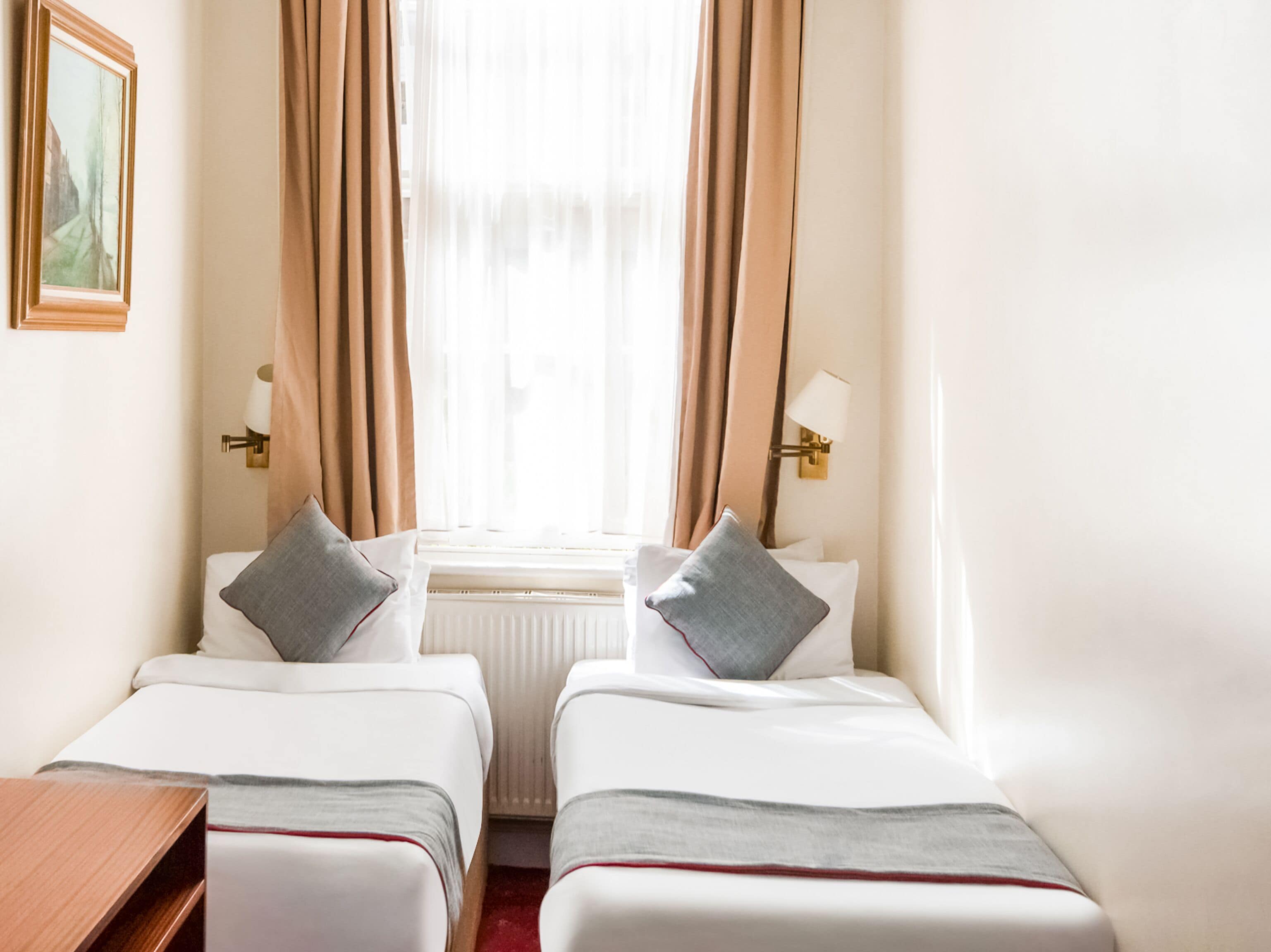 A hotel room with two single beds, a wooden nightstand, a radiator, and a window with curtains.