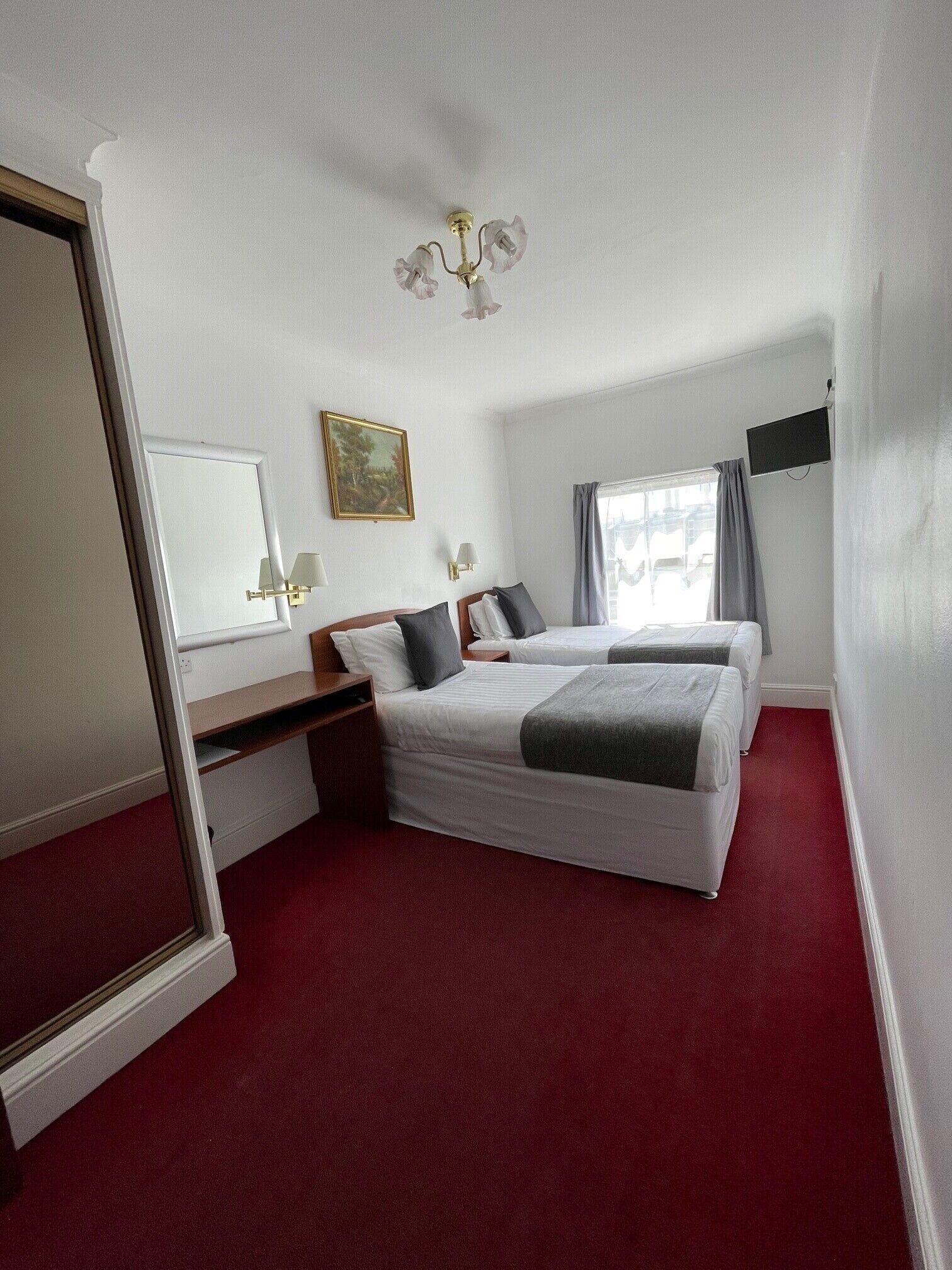 A hotel room with a large bed, a desk, a chair, a mirror, a painting, a television, and a window with curtains.