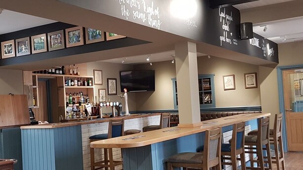 A bar area with a long counter, bar stools, and a wall-mounted TV. There are framed pictures on the wall and a shelf with bottles. - The Bowes Hotel (Hexham)