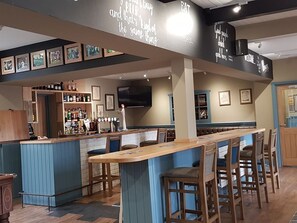 A bar area with a long counter, bar stools, and a wall-mounted TV. There are framed pictures on the wall and a shelf with bottles. - The Bowes Hotel (Hexham)