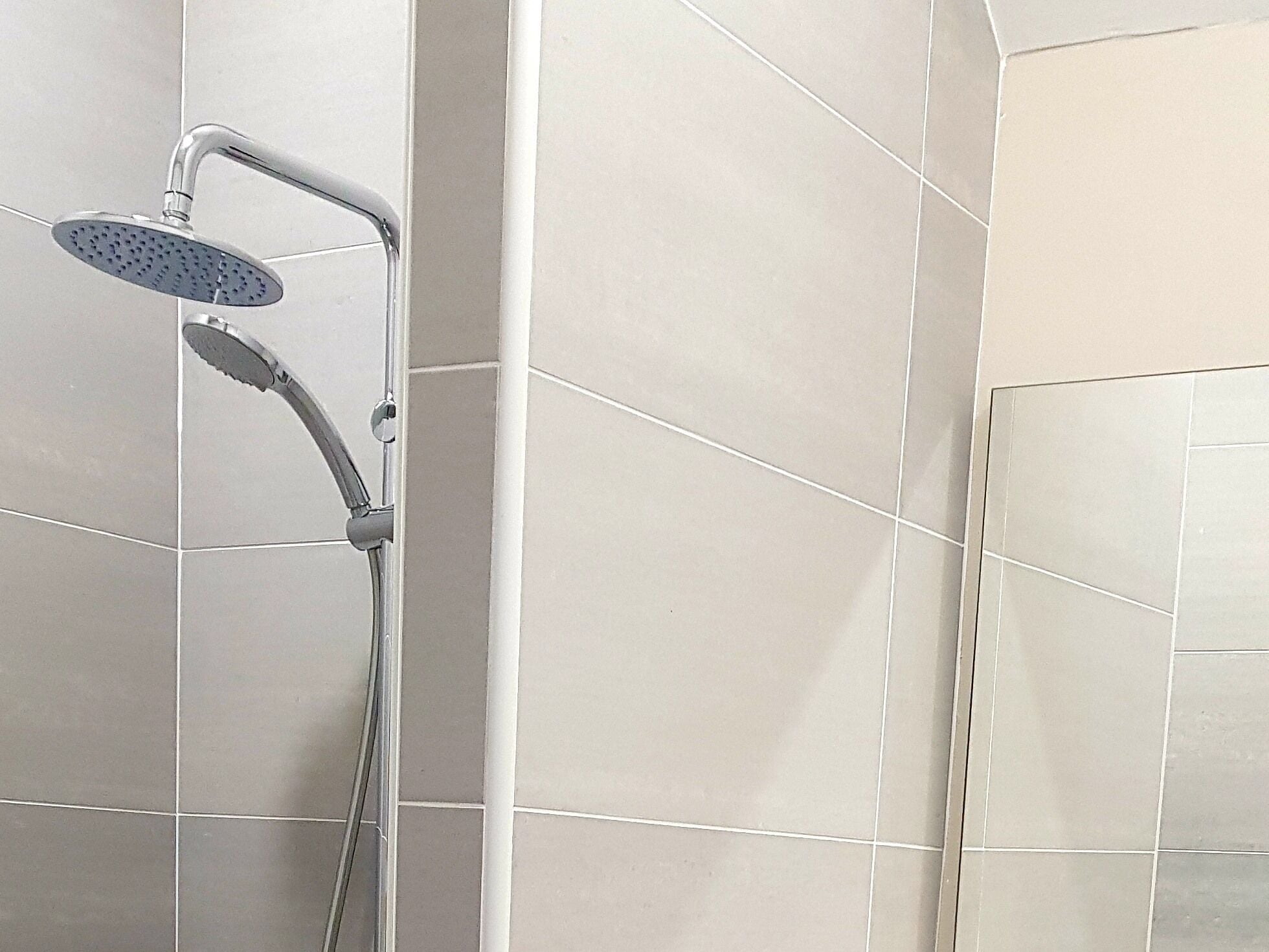 A shower with a handheld showerhead and a fixed overhead showerhead, mounted on a wall with large, light-colored tiles.