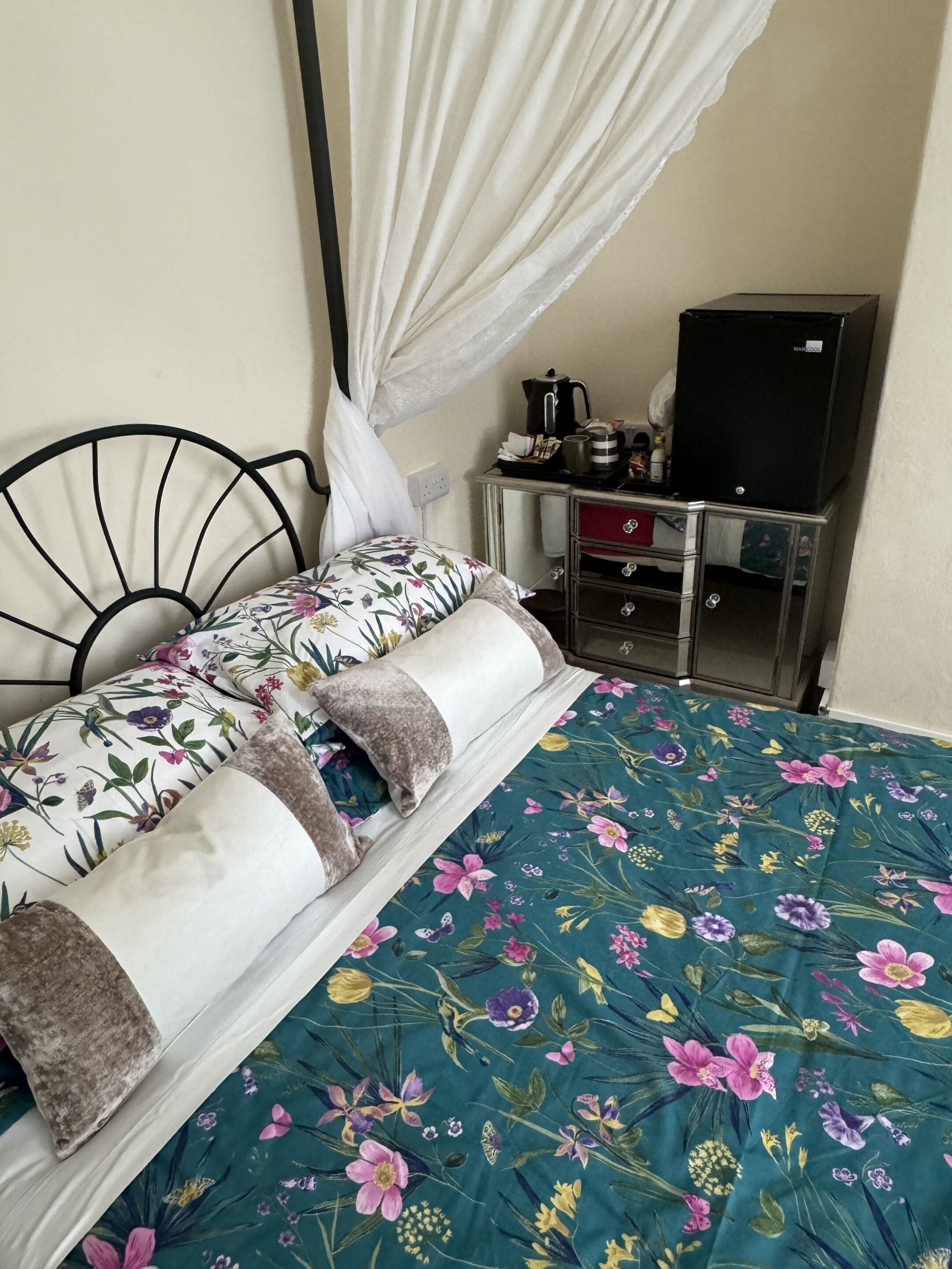 A bed with a floral bedspread and a canopy, a small bedside table with a kettle, and a black refrigerator.