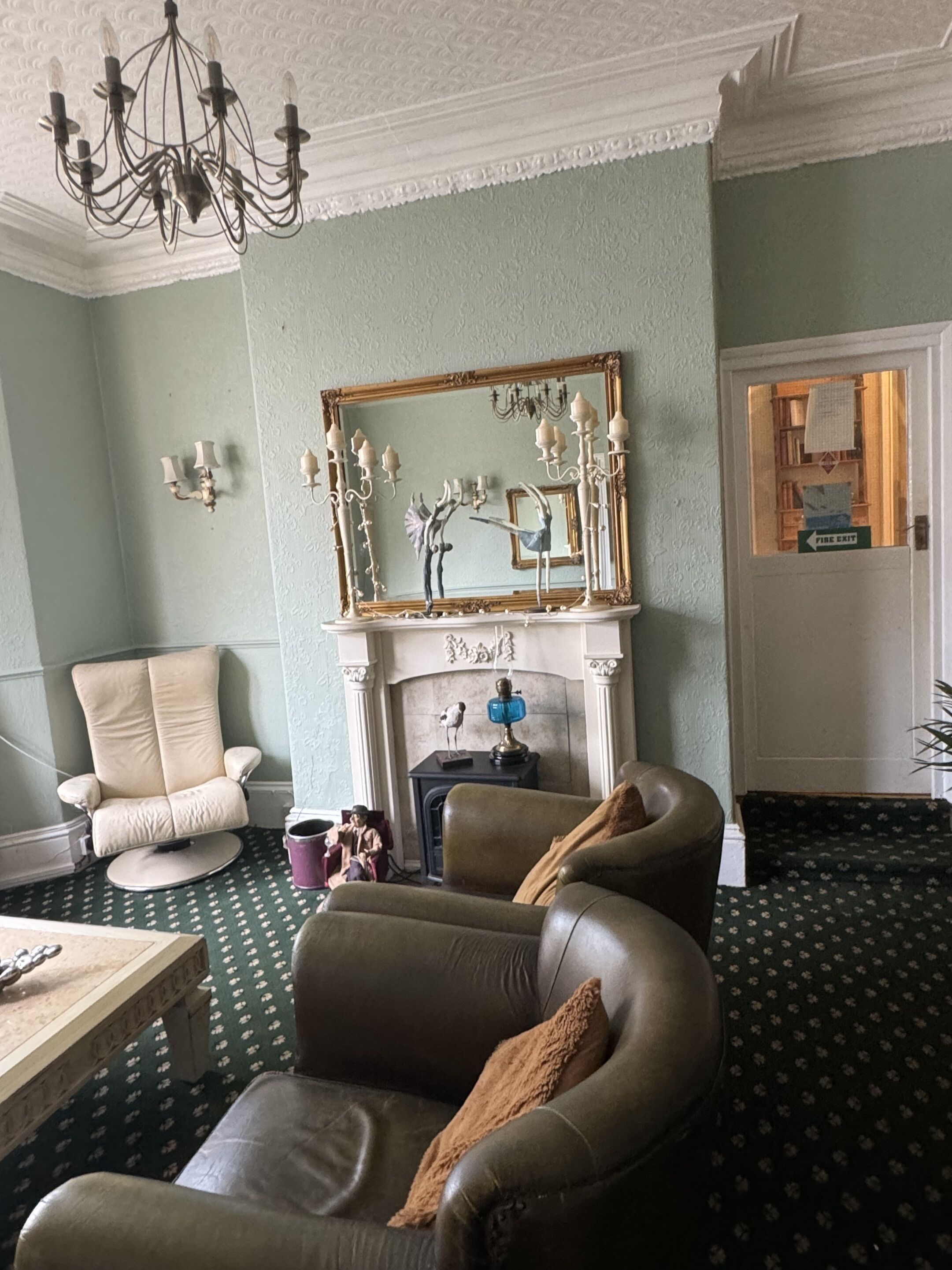 Un salon avec une cheminée, un lustre, un grand miroir et un panneau indiquant la sortie de secours.
