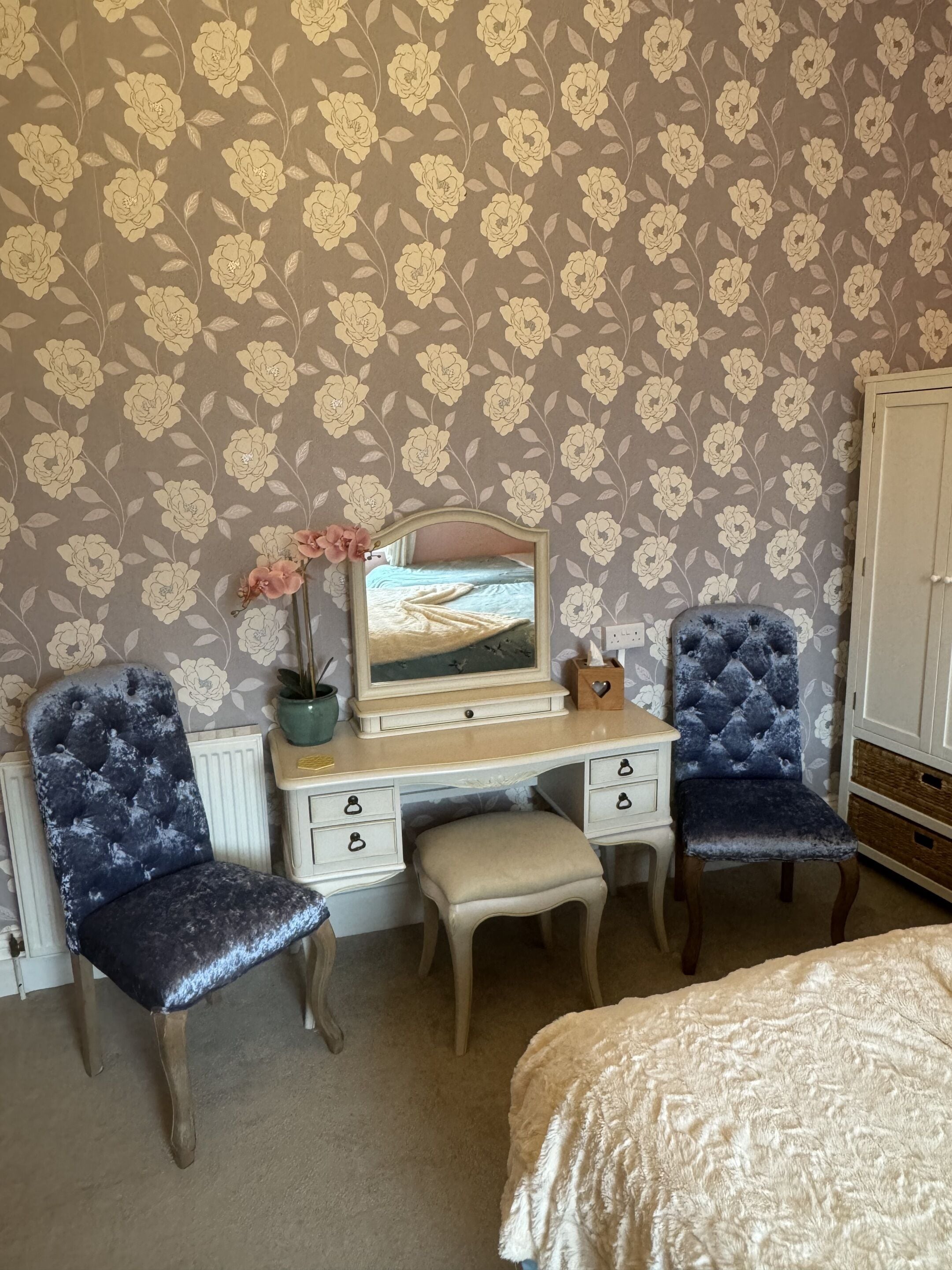 Une chambre avec un papier peint à motifs floraux, une commode, deux chaises, un lit et un miroir.