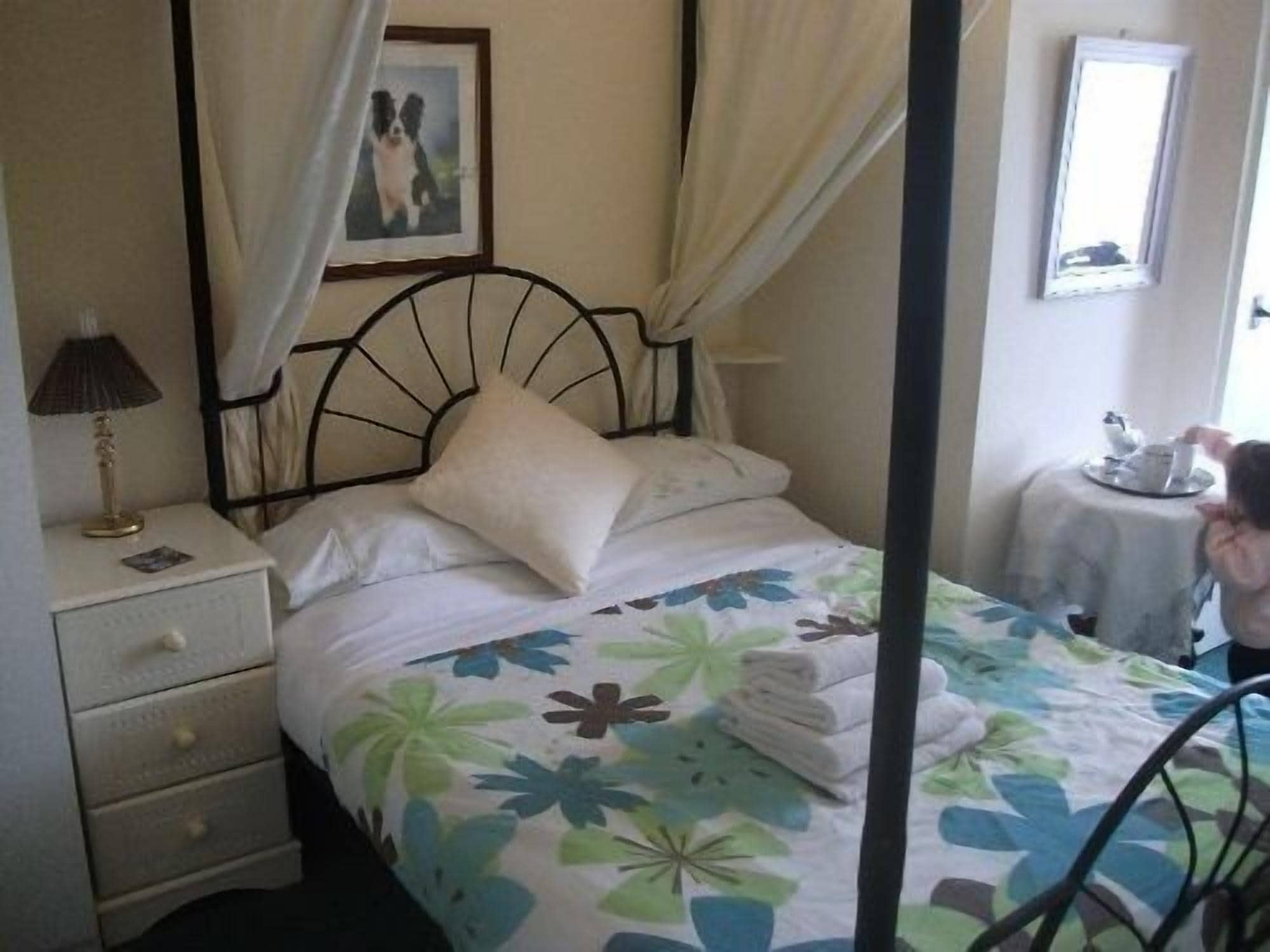 A four-poster bed with a floral bedspread, a bedside table with a lamp, and a framed picture on the wall.
