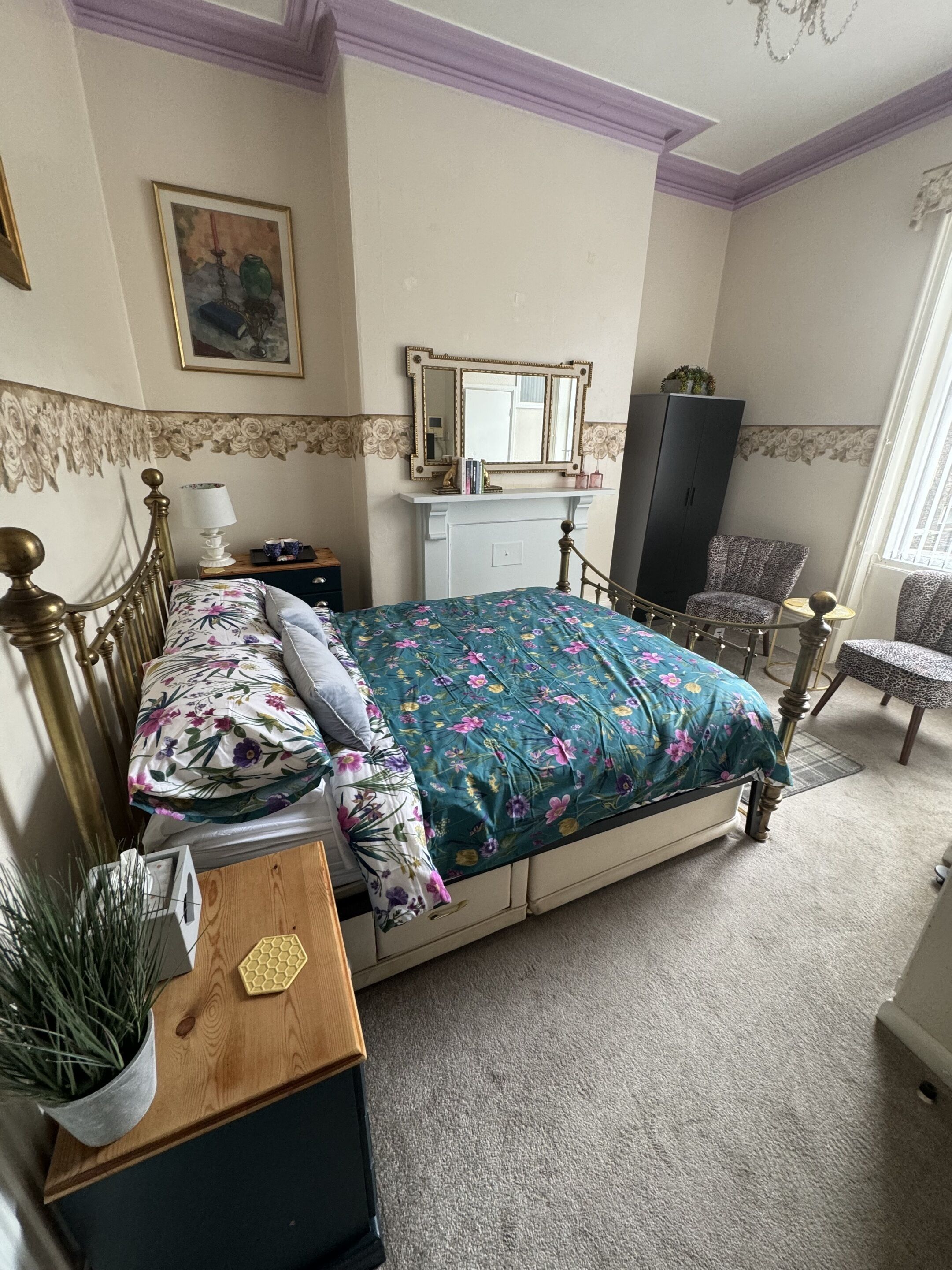 A bedroom with a floral bedspread, a wooden bedside table with a plant, a dresser, a chair, and a mirror above the dresser.