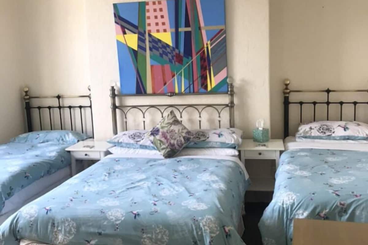 Ein Zimmer mit drei Betten, jedes mit einem floralen Überwurf, einem weißen Kopfteil und einem kleinen Nachttisch mit Lampe. Über dem mittleren Bett hängt ein farbenfrohes, abstraktes Gemälde.