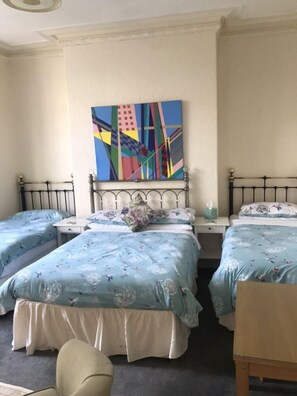 A room with three beds, each with a floral bedspread, a white headboard, and a small bedside table with a lamp. A colorful abstract painting hangs above the center bed. - Albemarle Hotel (Scarborough)