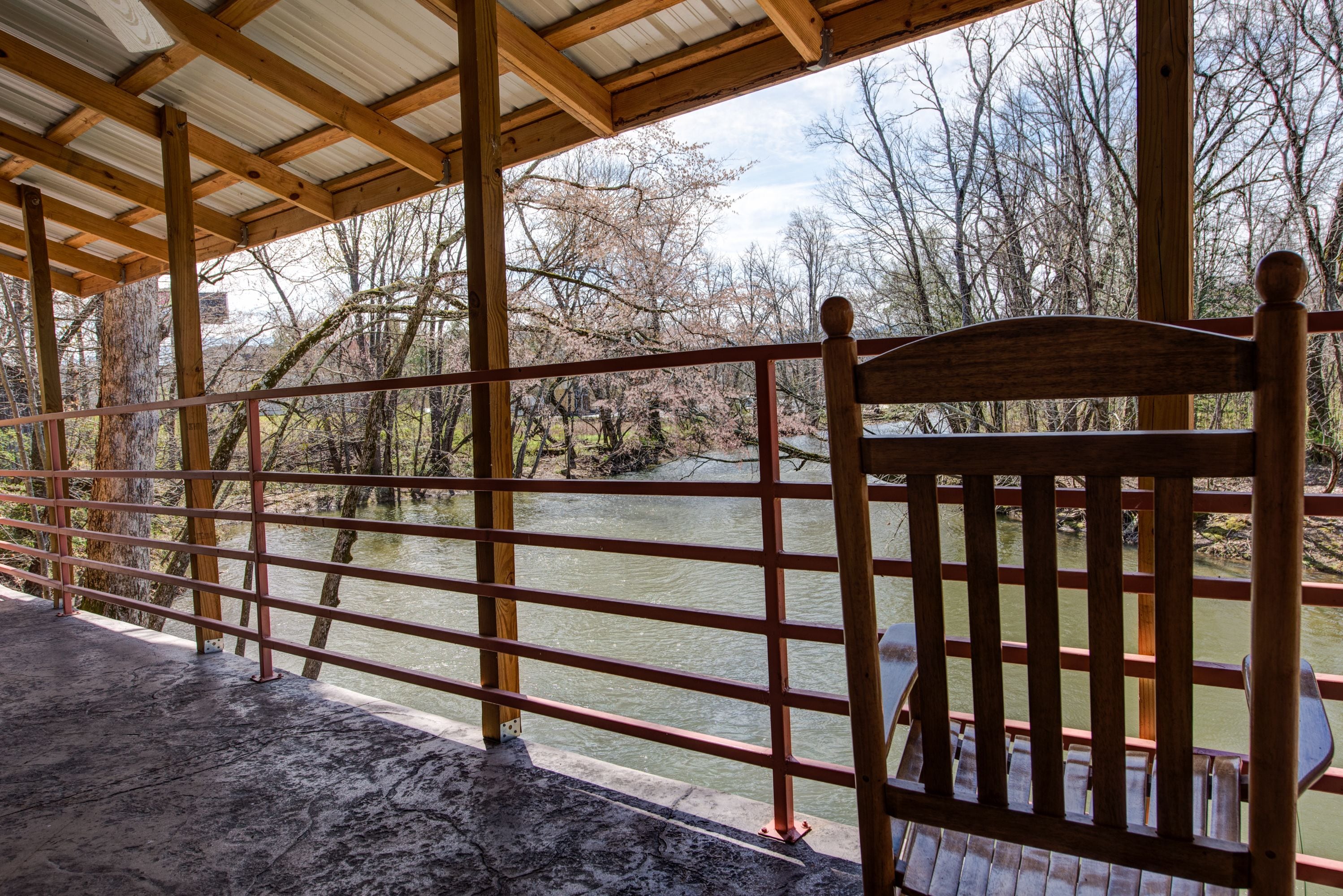 A covered outdoor area with a wooden bench and metal railing overlooking a body of water and trees.