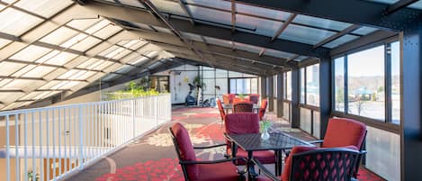 A covered outdoor seating area with a table and chairs, a patterned carpet, and large windows offering a view of the outdoors.