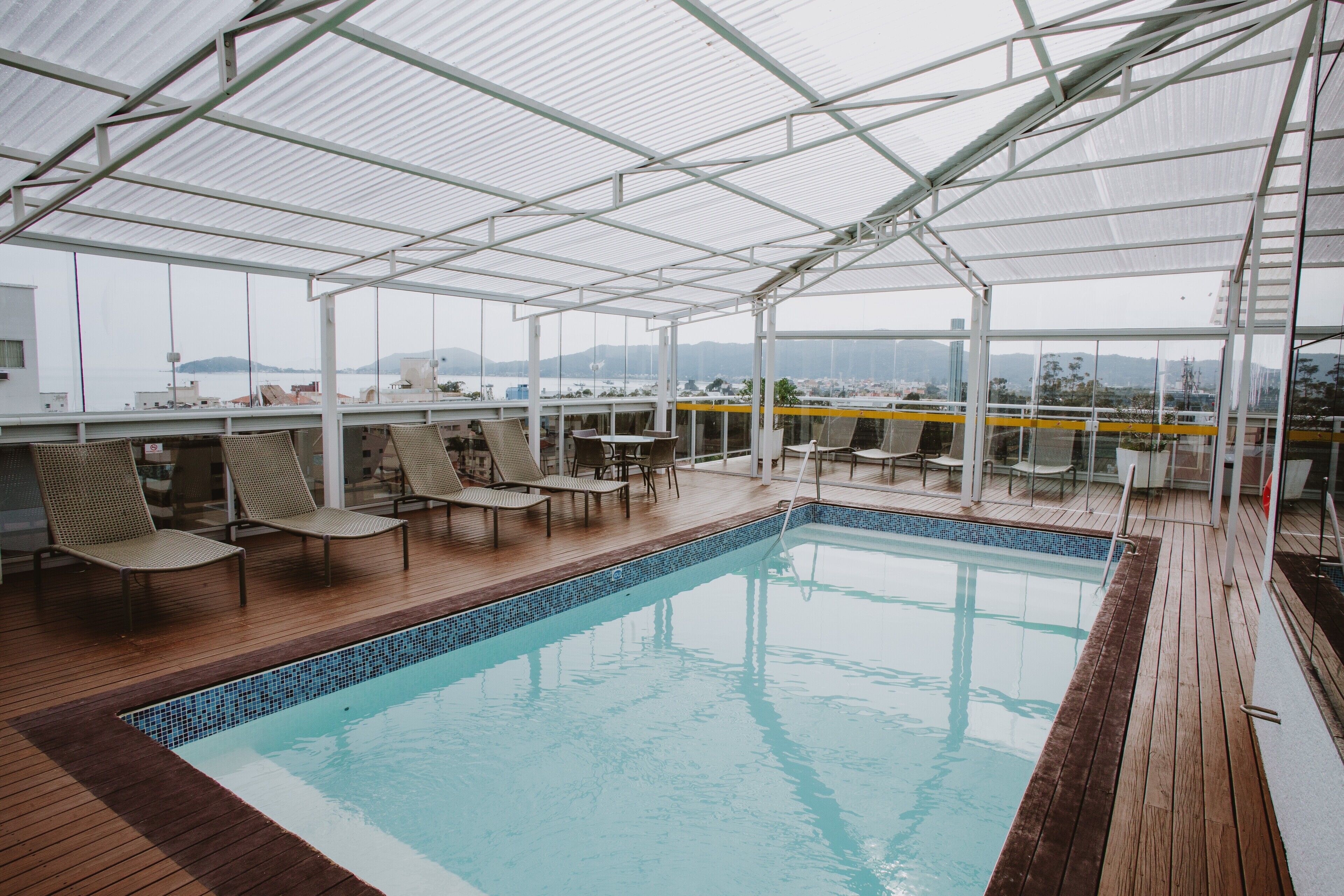 A covered swimming pool with lounge chairs and tables, surrounded by glass walls offering views of the city and hills.