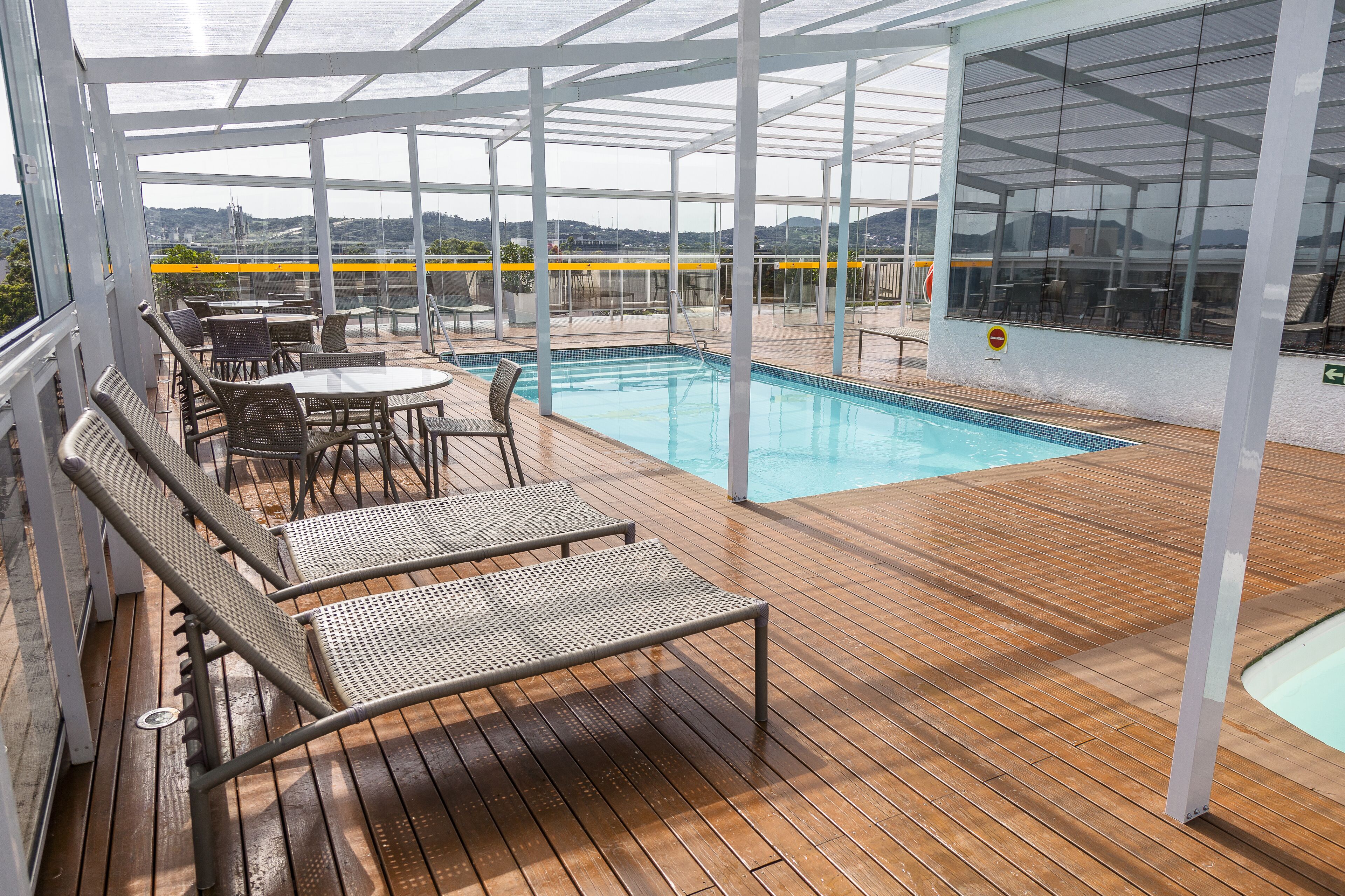A covered outdoor swimming pool area with lounge chairs and tables, surrounded by glass walls offering views of the landscape.