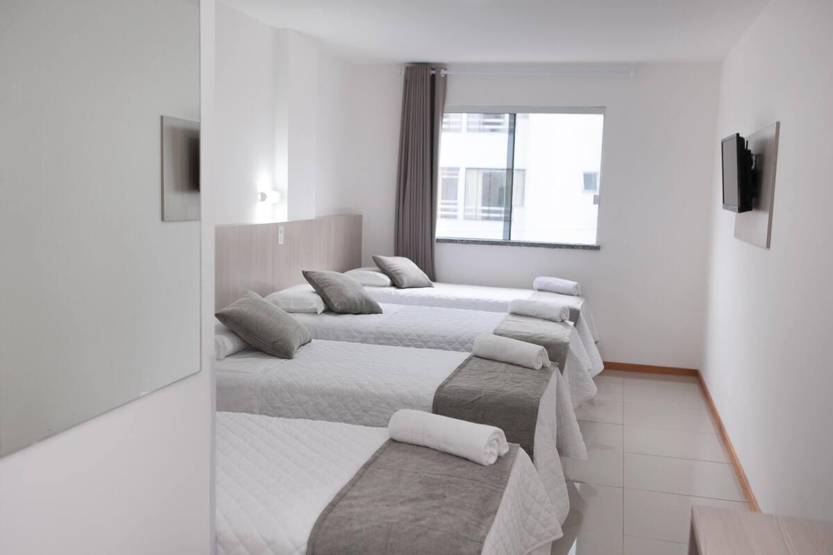 Ein Hotelzimmer mit vier nebeneinander stehenden Betten, einem großen Fenster mit Blick auf Gebäude, einem an der Wand montierten Flachbildfernseher und einem Holzboden.