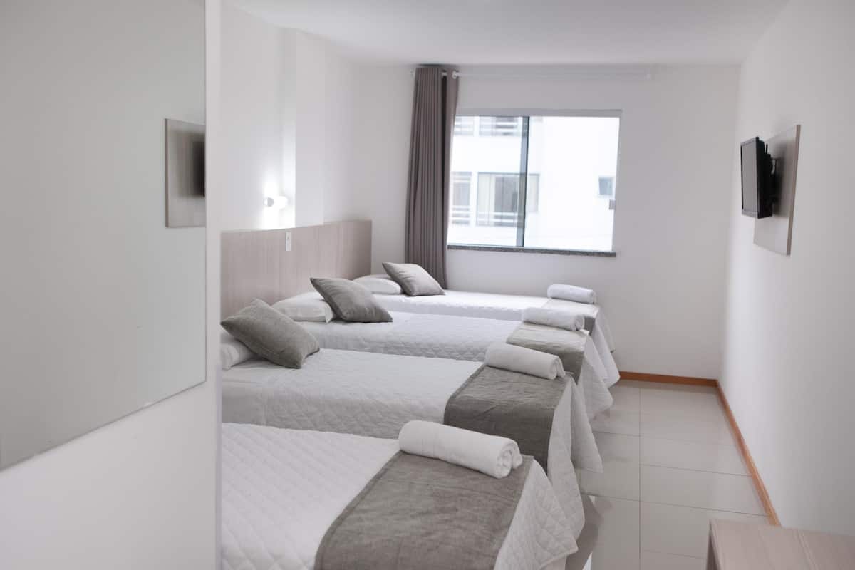Habitación de hotel con cuatro camas dispuestas en fila, un ventanal con vistas a edificios, un televisor de pantalla plana montado en la pared y suelo de madera.
