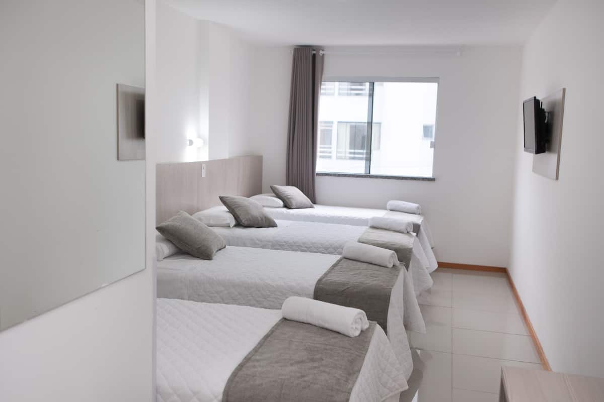 Ein Hotelzimmer mit vier nebeneinanderstehenden Betten, einem großen Fenster mit Vorhängen, einem an der Wand befestigten Flachbildfernseher und einem Holzboden.