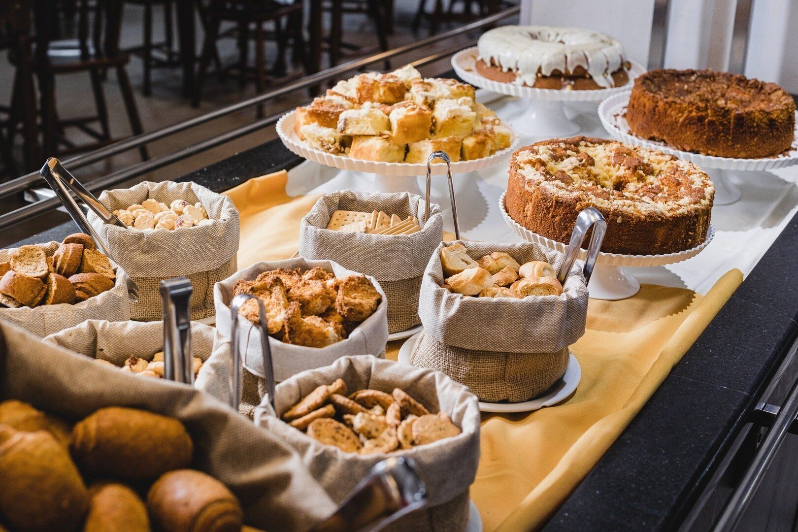 Uma mesa de buffet com diversos produtos de panificação e doces, incluindo pão, bolos e biscoitos, dispostos sobre uma toalha de mesa.