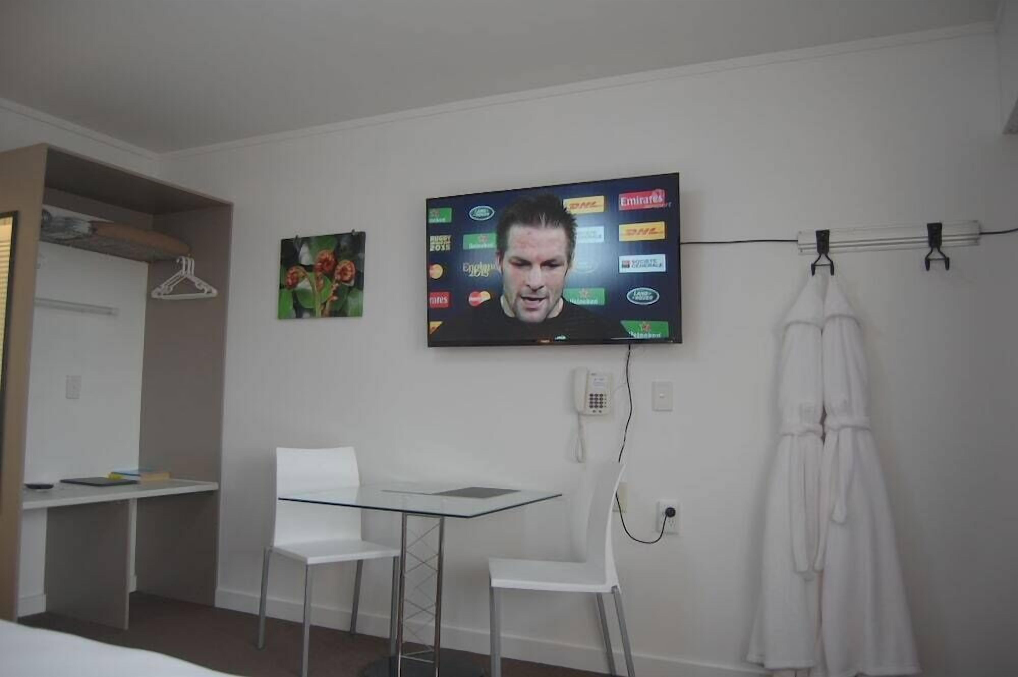 A room with a TV mounted on the wall displaying various logos, a white desk with chairs, a coat rack with a robe, and a small painting.