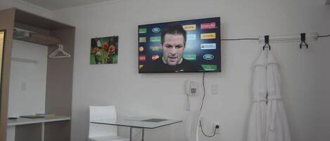 A room with a TV mounted on the wall displaying various logos, a white desk with chairs, a coat rack with a robe, and a small painting.