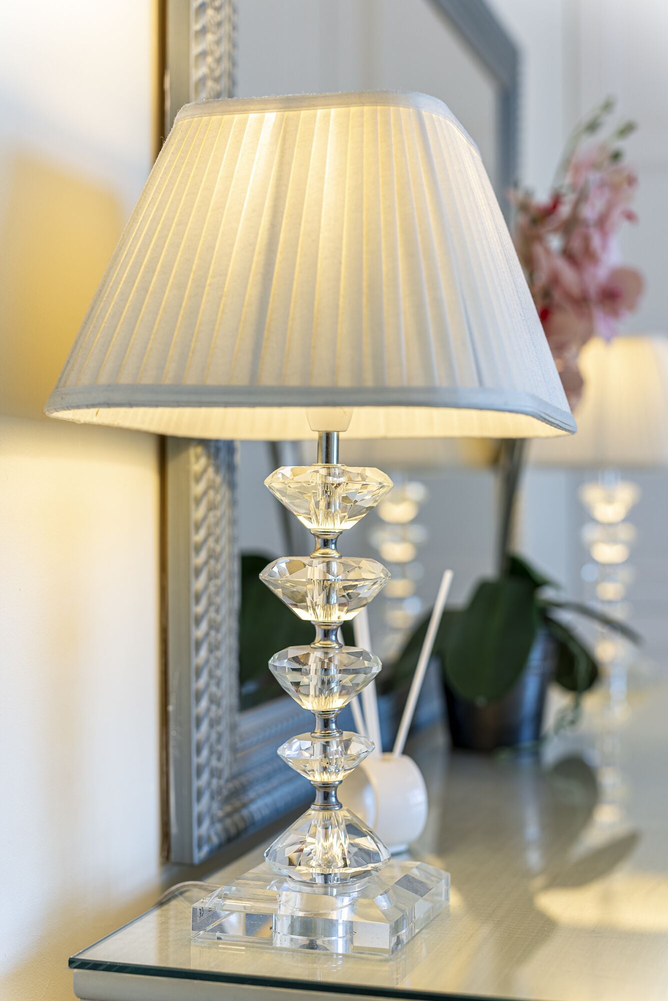 Uma luminária moderna com cúpula plissada e base de cristal sobre uma superfície espelhada.