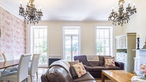 A spacious living room with a brown leather sofa, a wooden coffee table, and a dining area with a white table and chairs. Large windows allow natural light to fill the room.