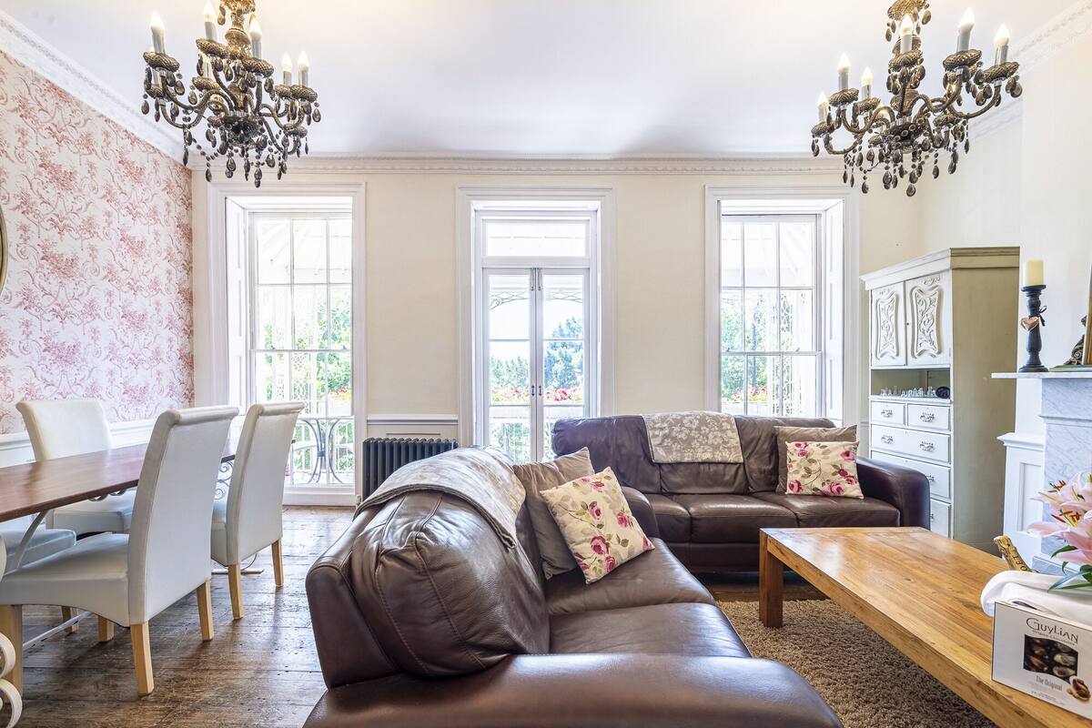 Een ruime woonkamer met een bruine leren bank, een houten salontafel en een eethoek met een witte tafel en stoelen. Grote ramen zorgen voor veel natuurlijk licht in de kamer.