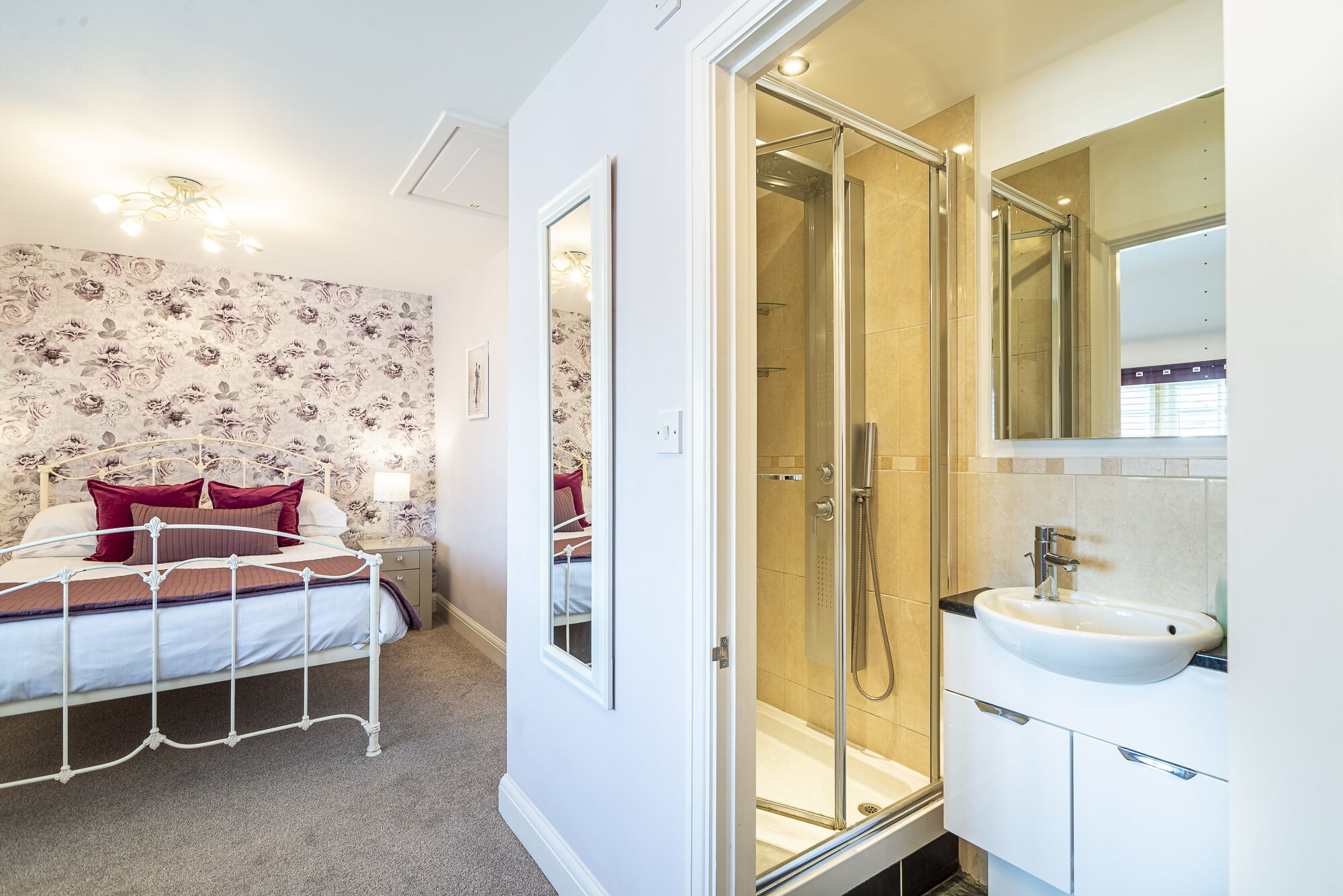 A bedroom with a bed, a bedside table, and a nightstand. There is a shower area with a glass door and a sink with a mirror above it.