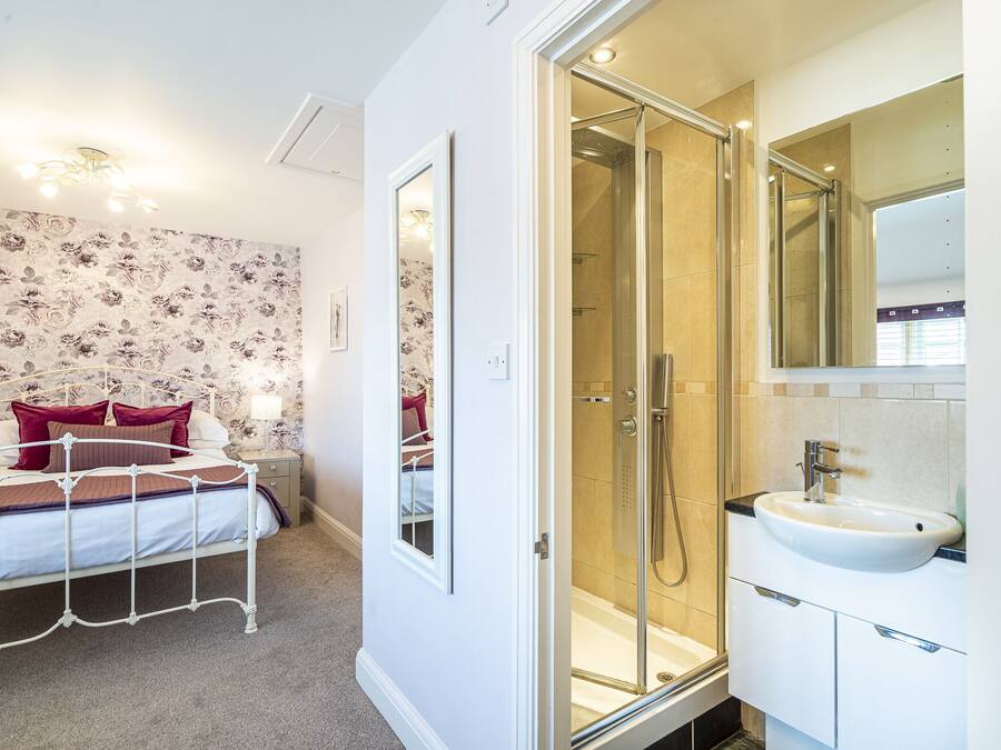 A bedroom with a bed, a bedside table, and a nightstand. There is a shower area with a glass door and a sink with a mirror above it.