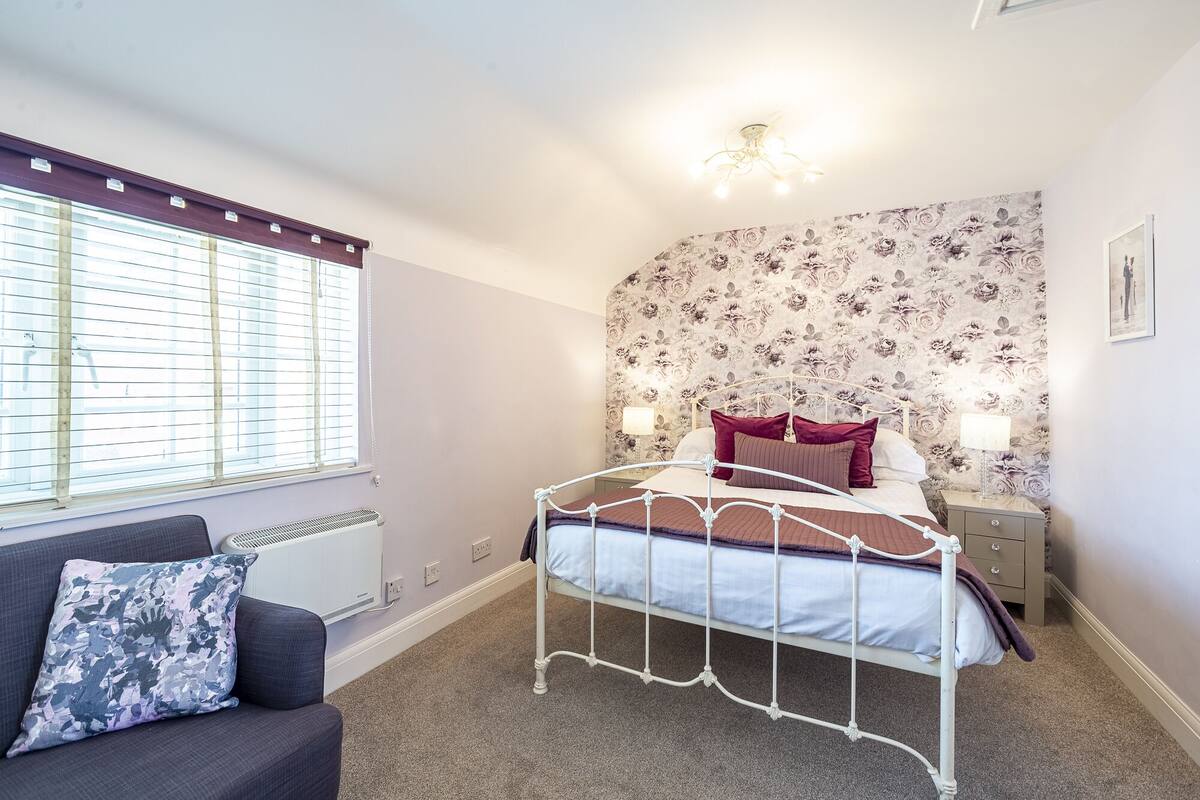 Een slaapkamer met een bed, nachtkastjes, een radiator, een raam met jaloezieën en een wand met bloemenbehang.