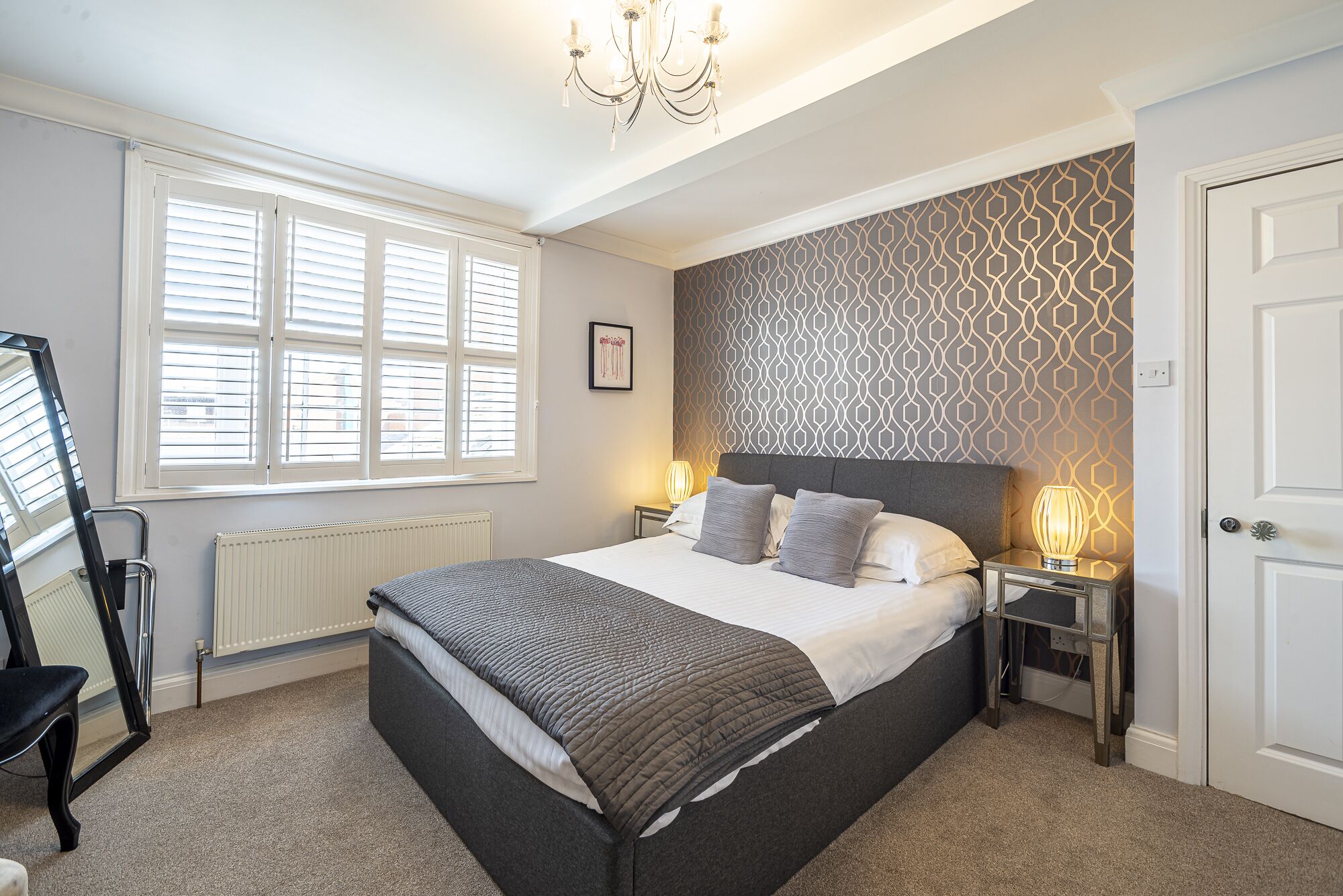 A bedroom with a bed, two bedside tables, a radiator, a window with blinds, a door, and a chandelier.