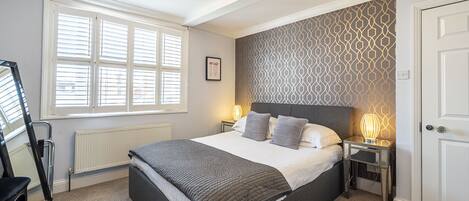 A bedroom with a bed, two bedside tables, a radiator, a window with blinds, a door, and a chandelier.