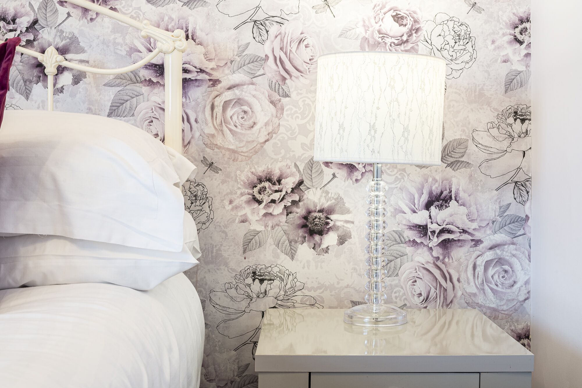 A bedroom with a floral wallpaper, a white bed, and a bedside table with a lamp.
