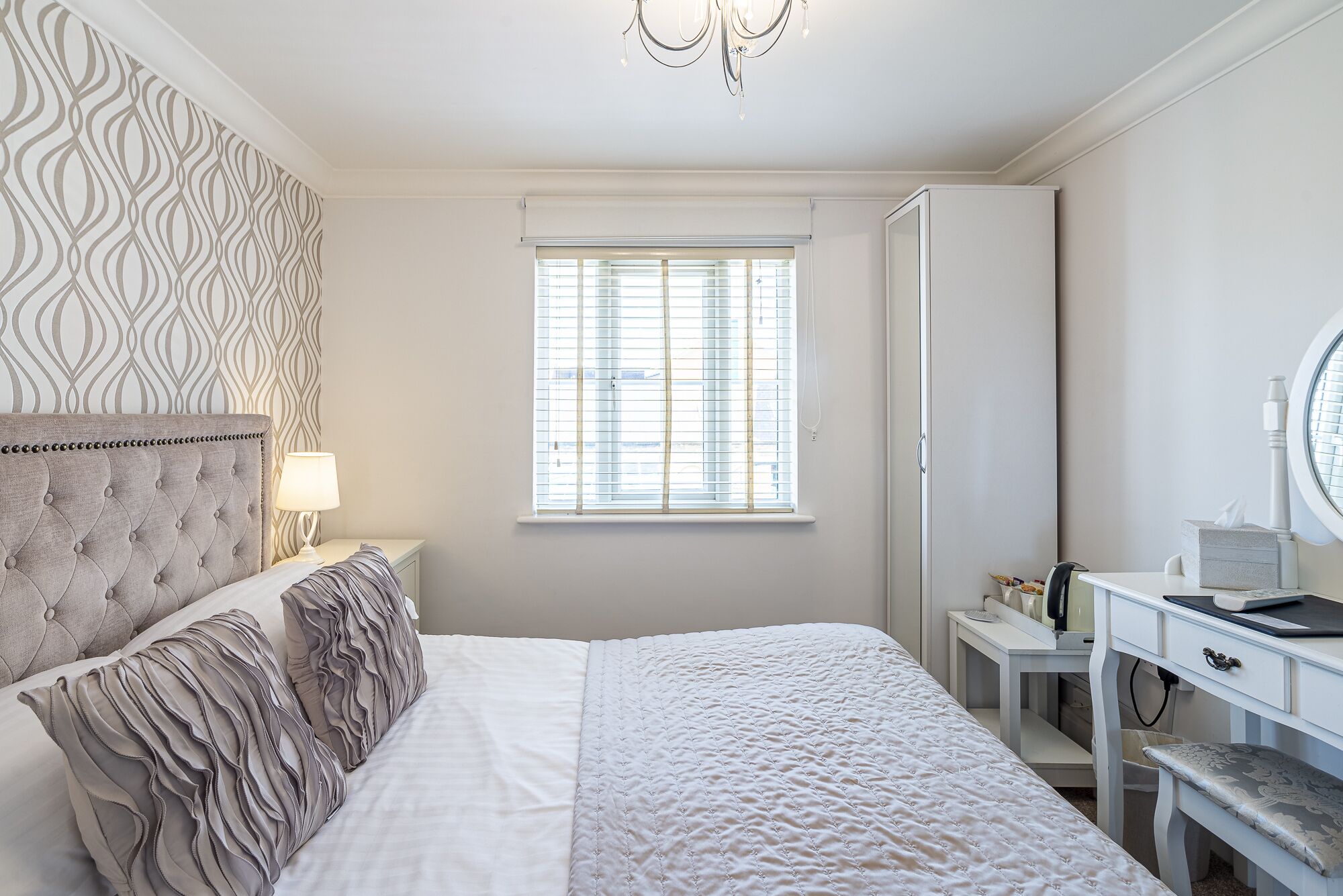 Un dormitorio con cama, una mesita de noche con lámpara, una ventana con persianas, un escritorio con silla y un espejo.