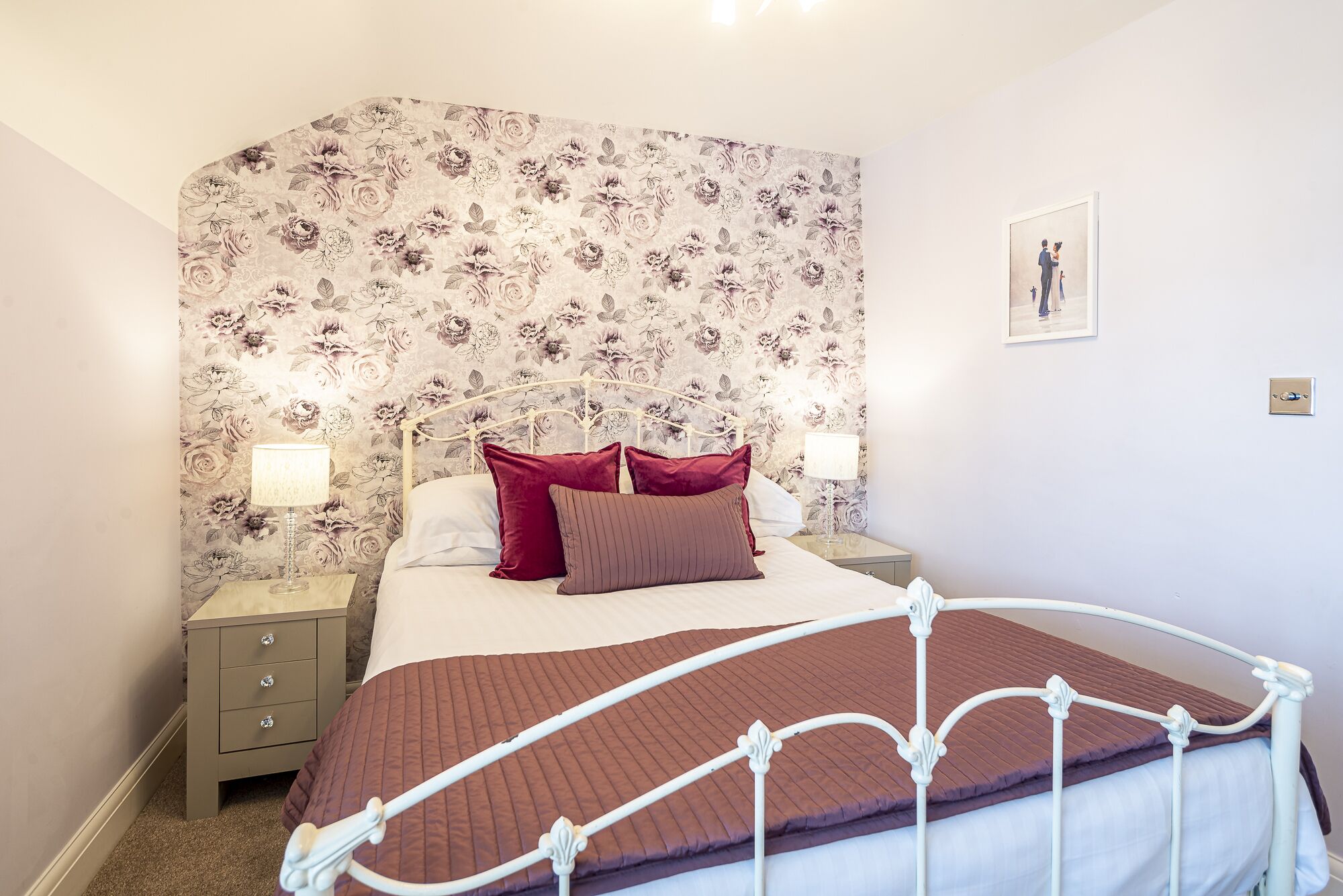 Un dormitorio con papel tapiz floral, estructura de cama de metal blanco y colcha marrón.