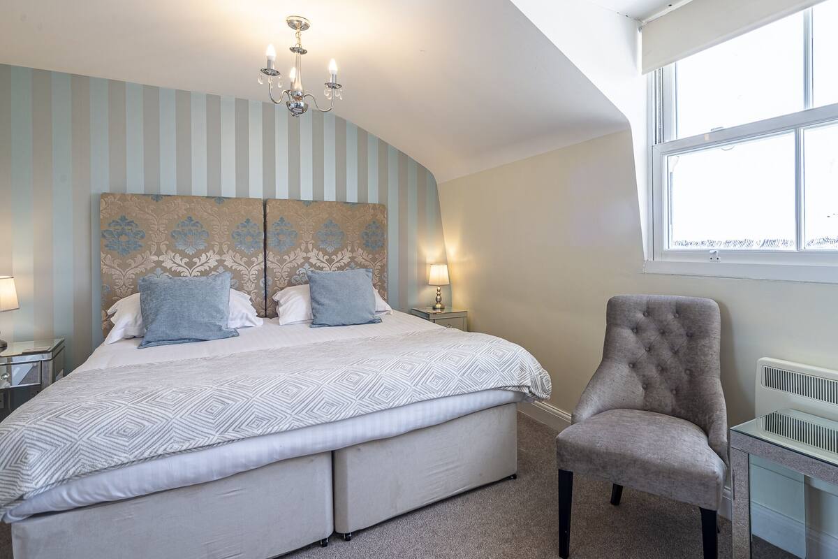 Een slaapkamer met een groot bed, een stoel, een nachtkastje, een lamp en een raam.