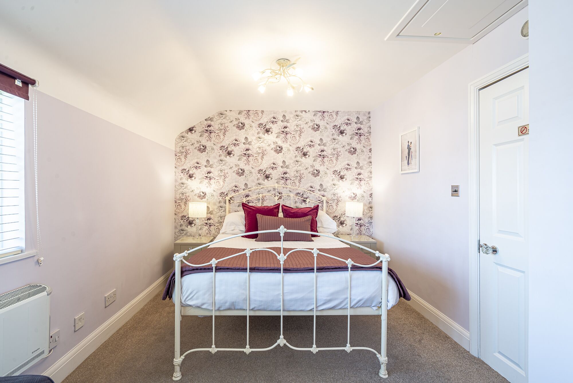 Une chambre avec un lit simple, un papier peint à motifs floraux, un luminaire au plafond et une porte blanche.