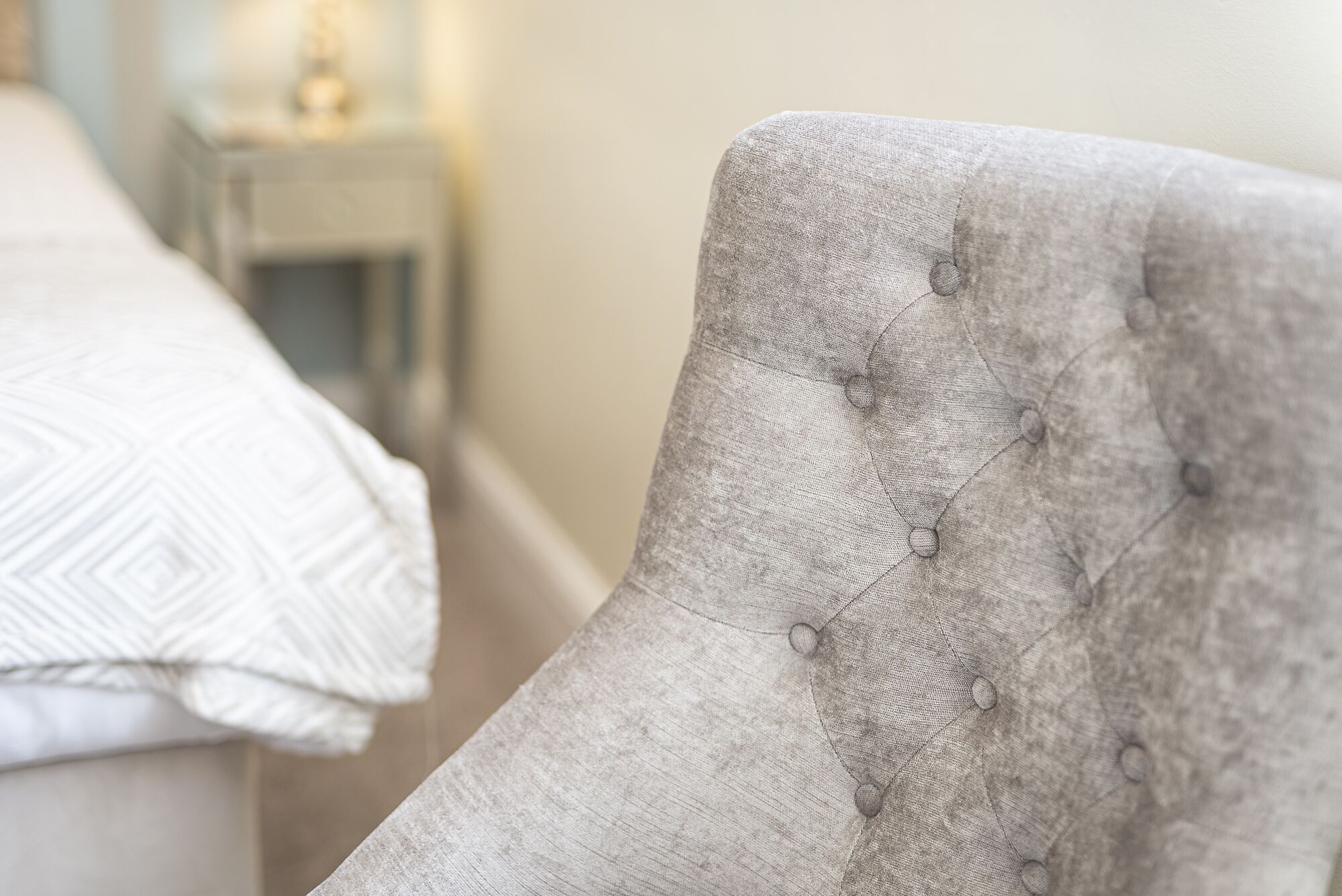A tufted armchair with a buttoned backrest, positioned next to a bed with a patterned white duvet.