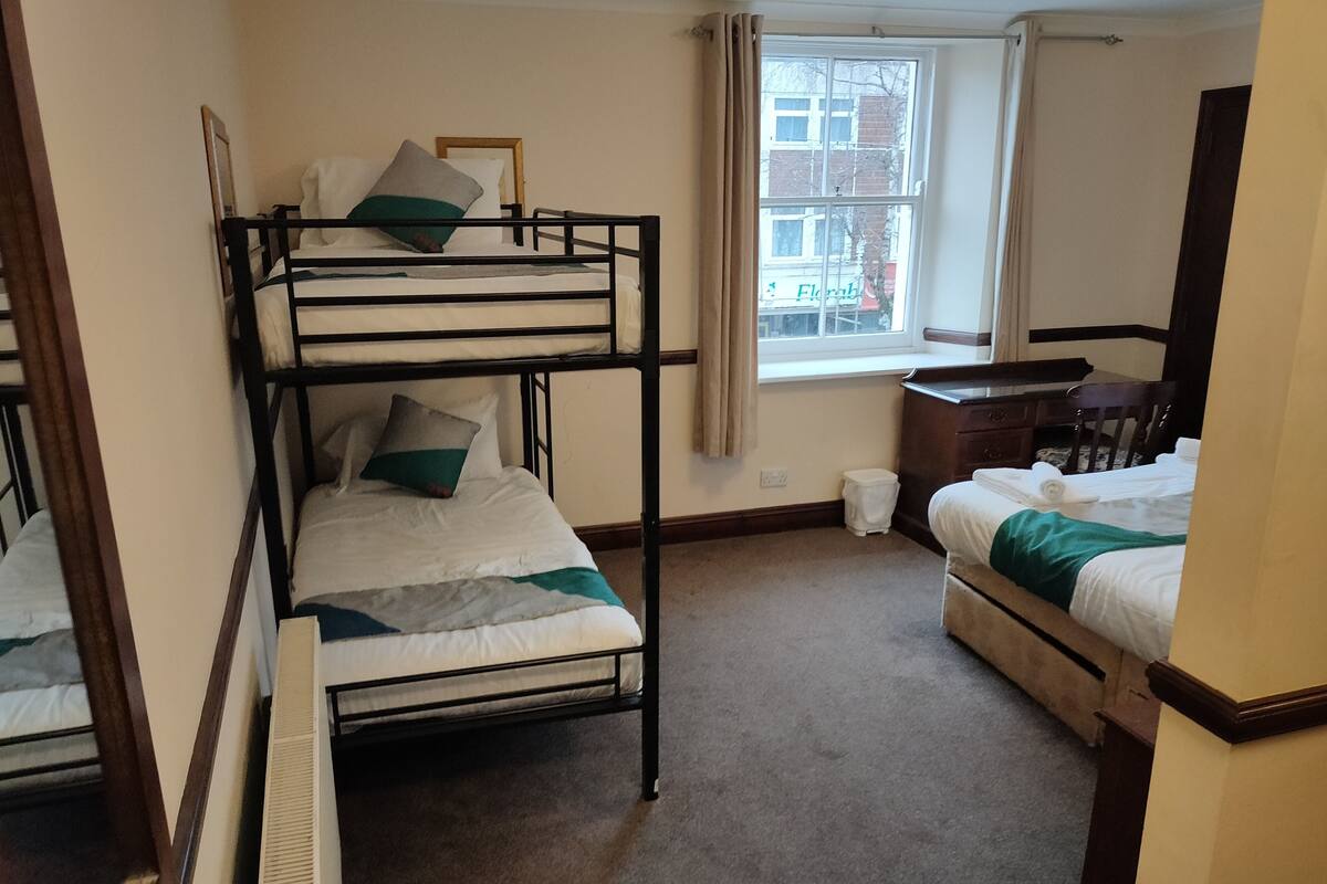 A dormitory room with bunk beds, a desk, a chair, and a window with a view of a building outside.