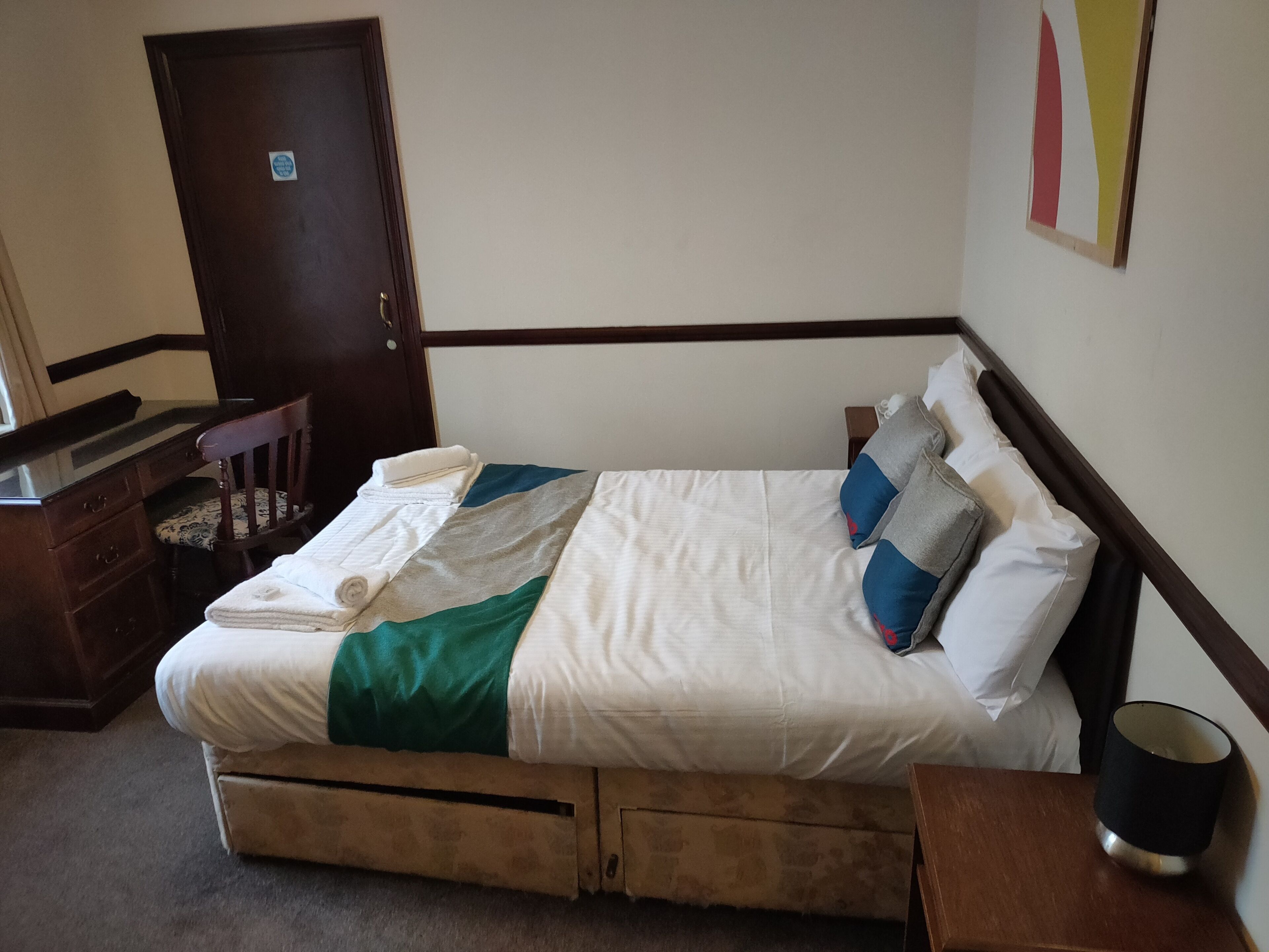A neatly made bed with pillows and a blanket, a wooden desk with drawers, a chair, and a door.