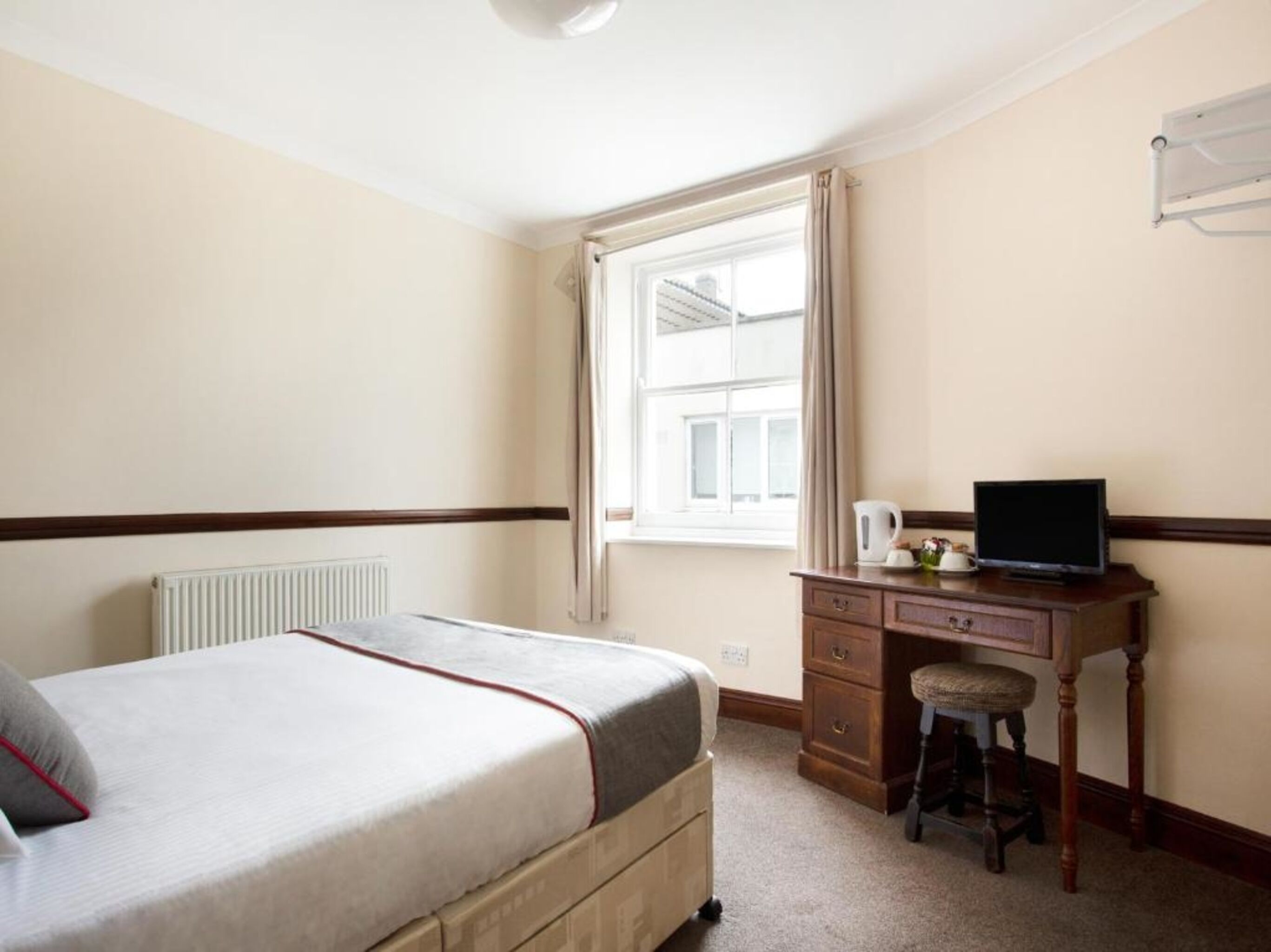 A bedroom with a bed, a desk with a computer, a radiator, and a window with curtains.