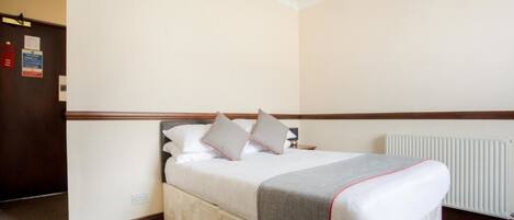 A hotel room with a bed, a radiator, and a door with a fire alarm.