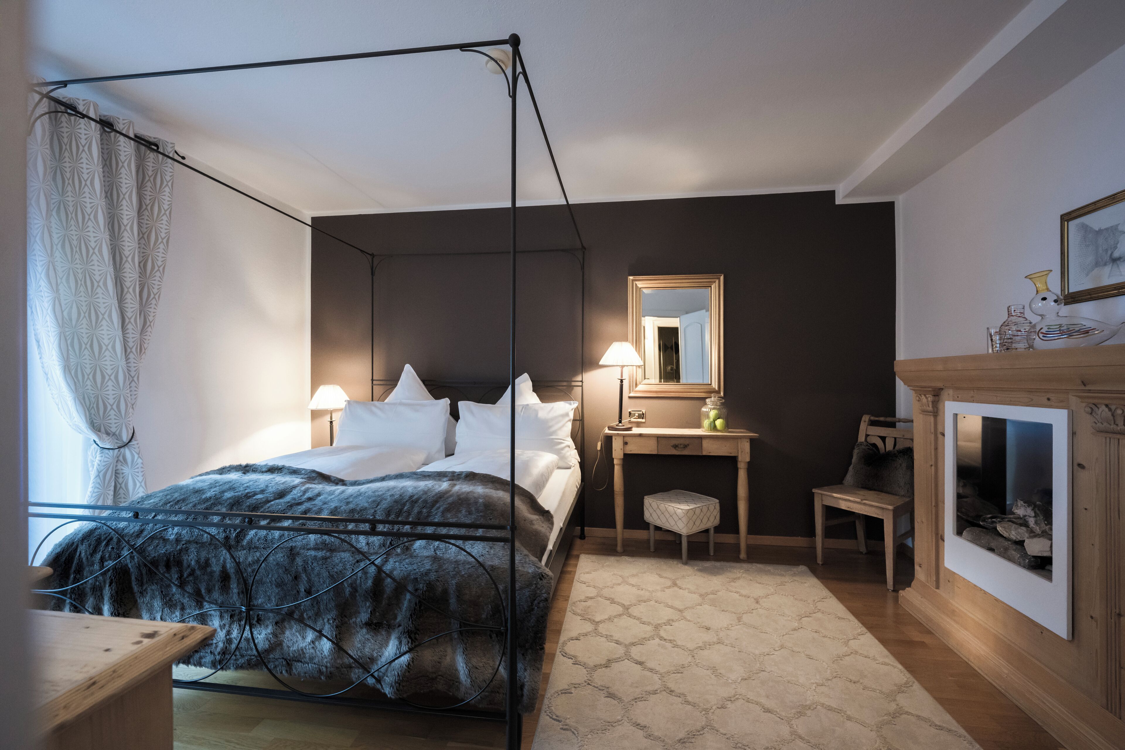 A bedroom with a four-poster bed, a bedside table with a lamp, a chair, a small stool, and a fireplace.